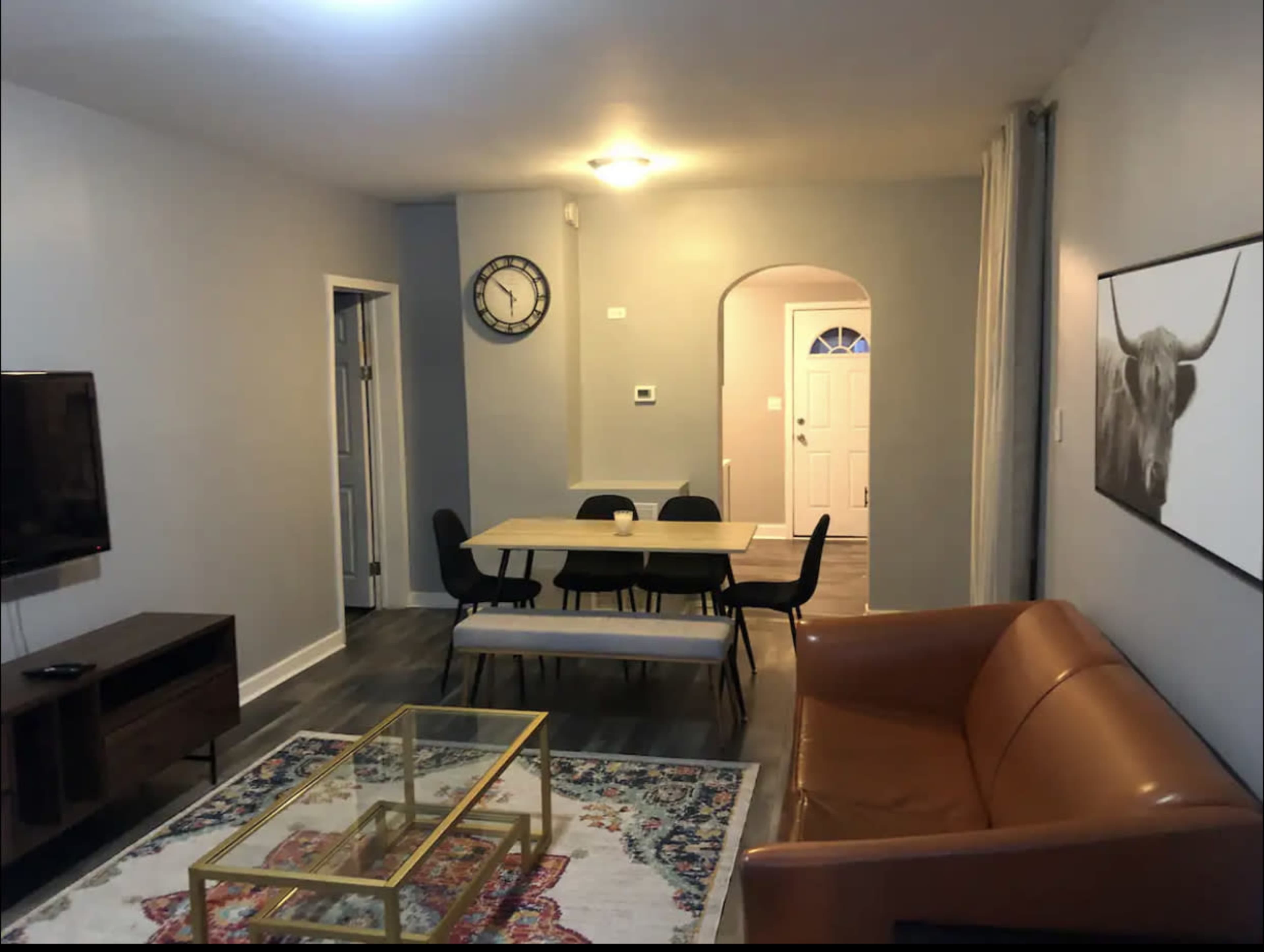 A modern living room features a brown leather sofa, a glass coffee table on a patterned rug, a dining table, and a wall-mounted television.
