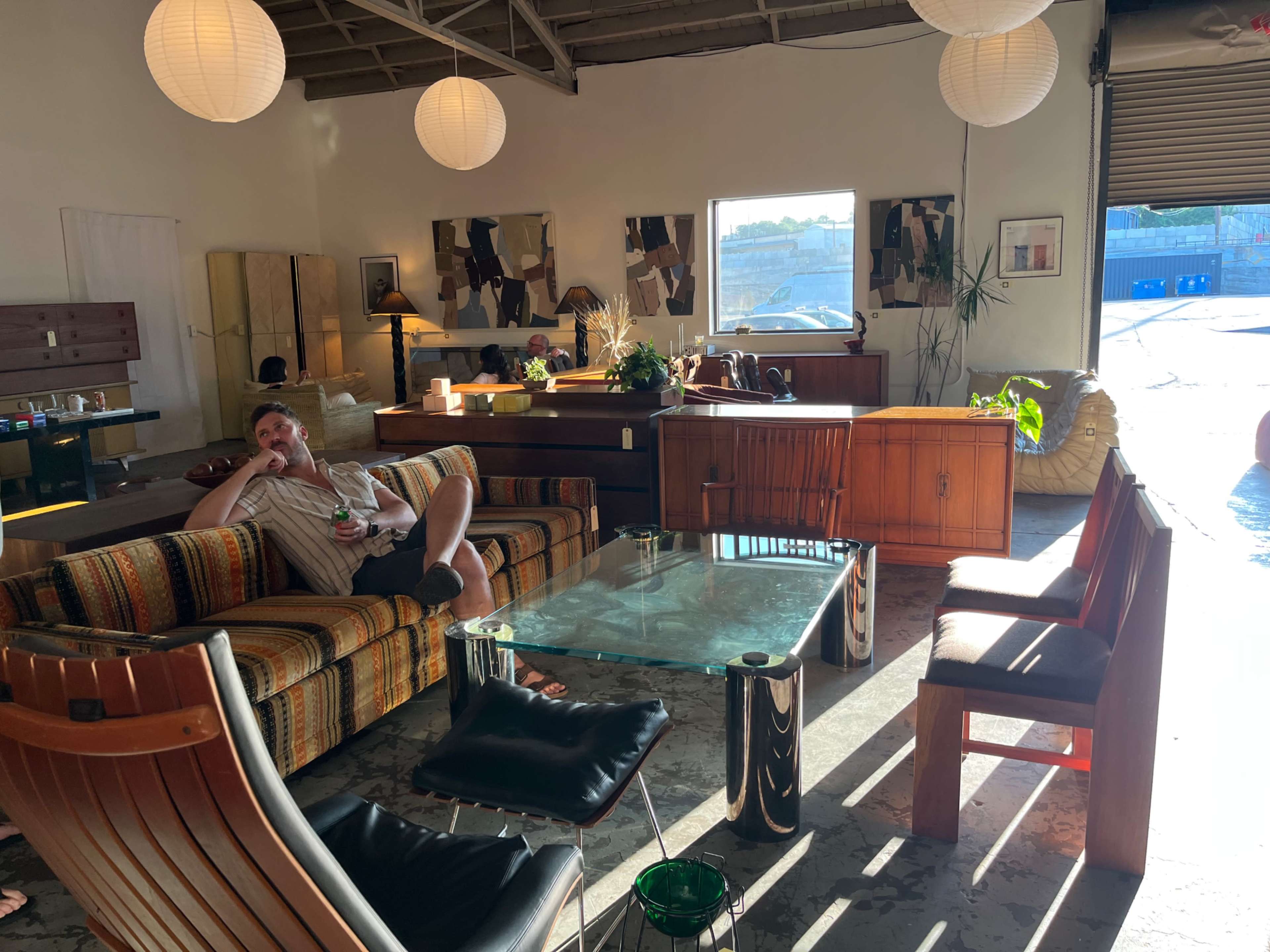 A man sits on a patterned sofa in a well-lit furniture showroom, surrounded by various wooden furniture pieces and decorative items.