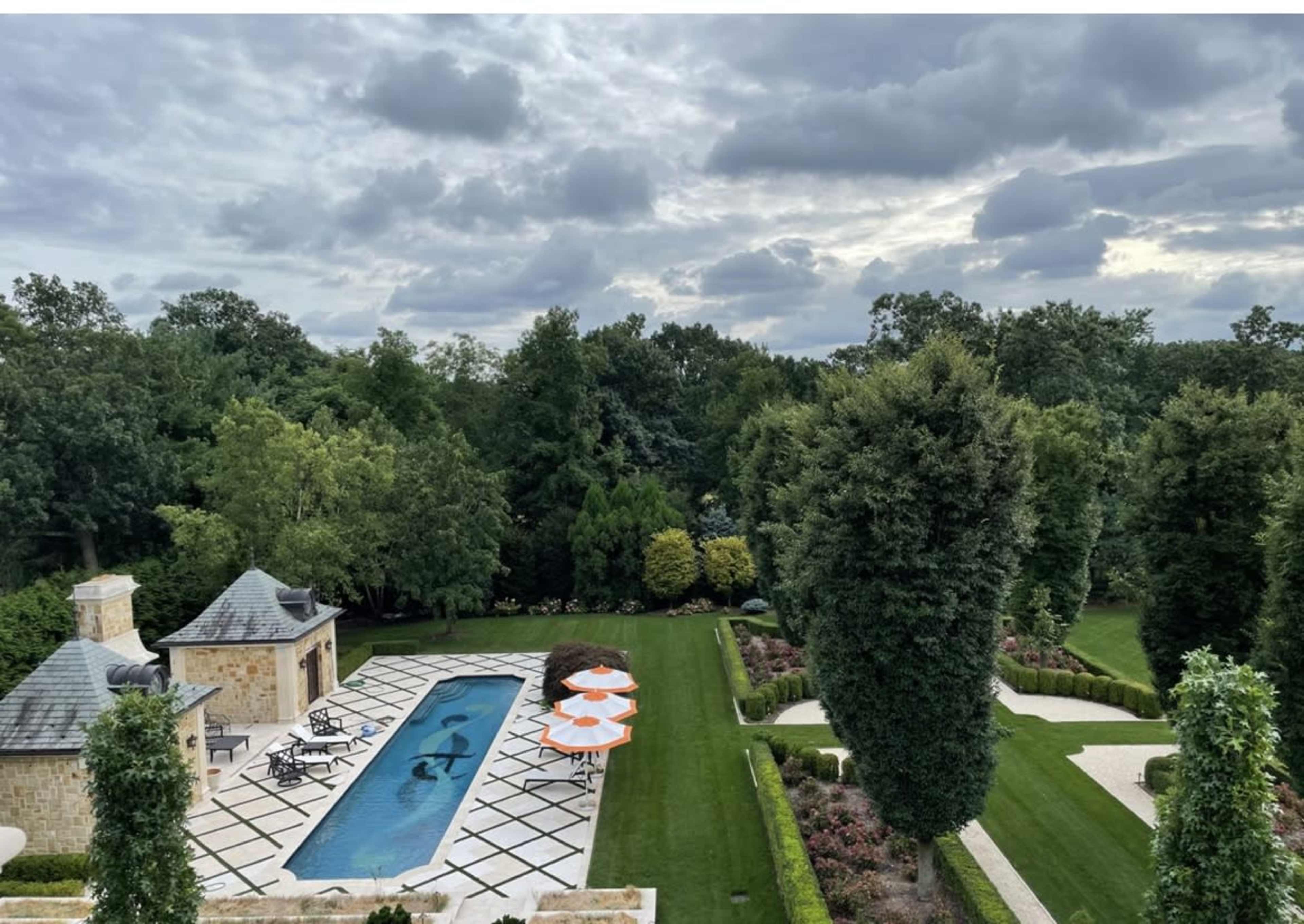 A view from above shows a rectangular swimming pool surrounded by neatly arranged lounge chairs and umbrellas, bordered by well-maintained gardens and trees.