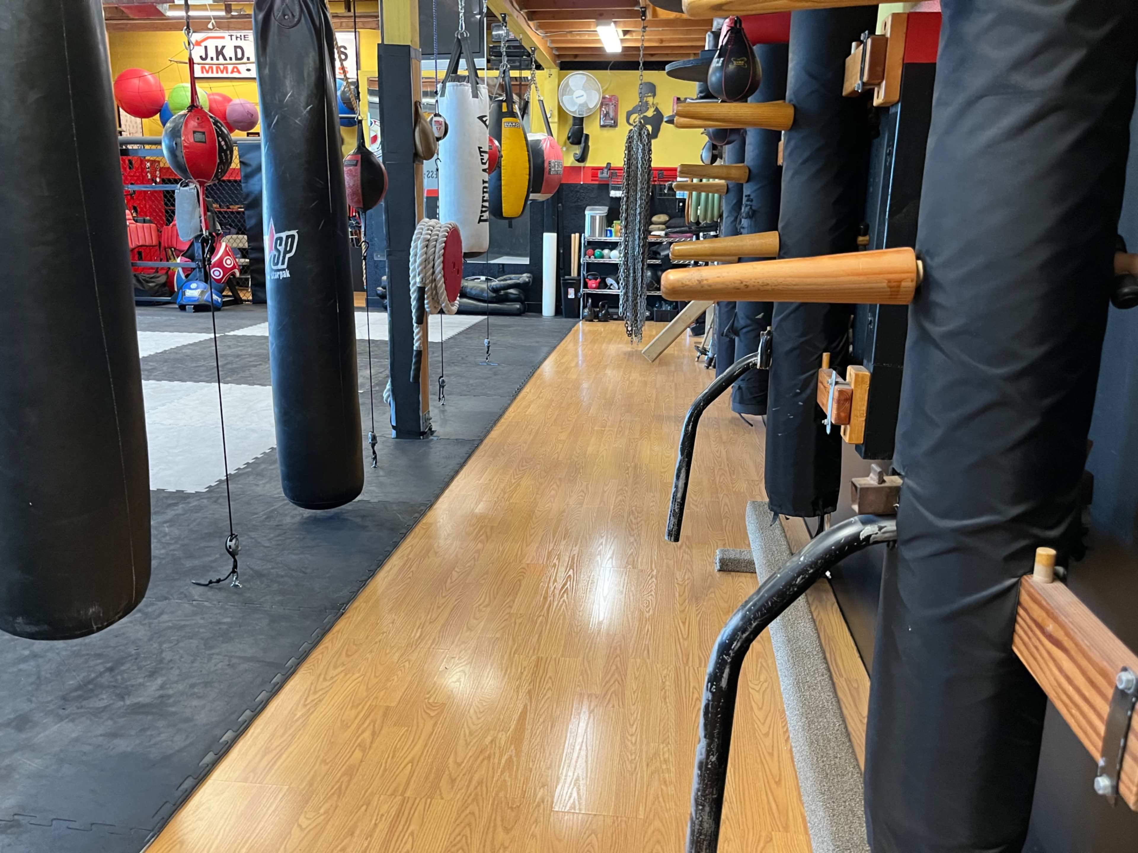 The image shows a training area in a gym with various hanging punching bags, wall-mounted equipment, and a wooden floor.