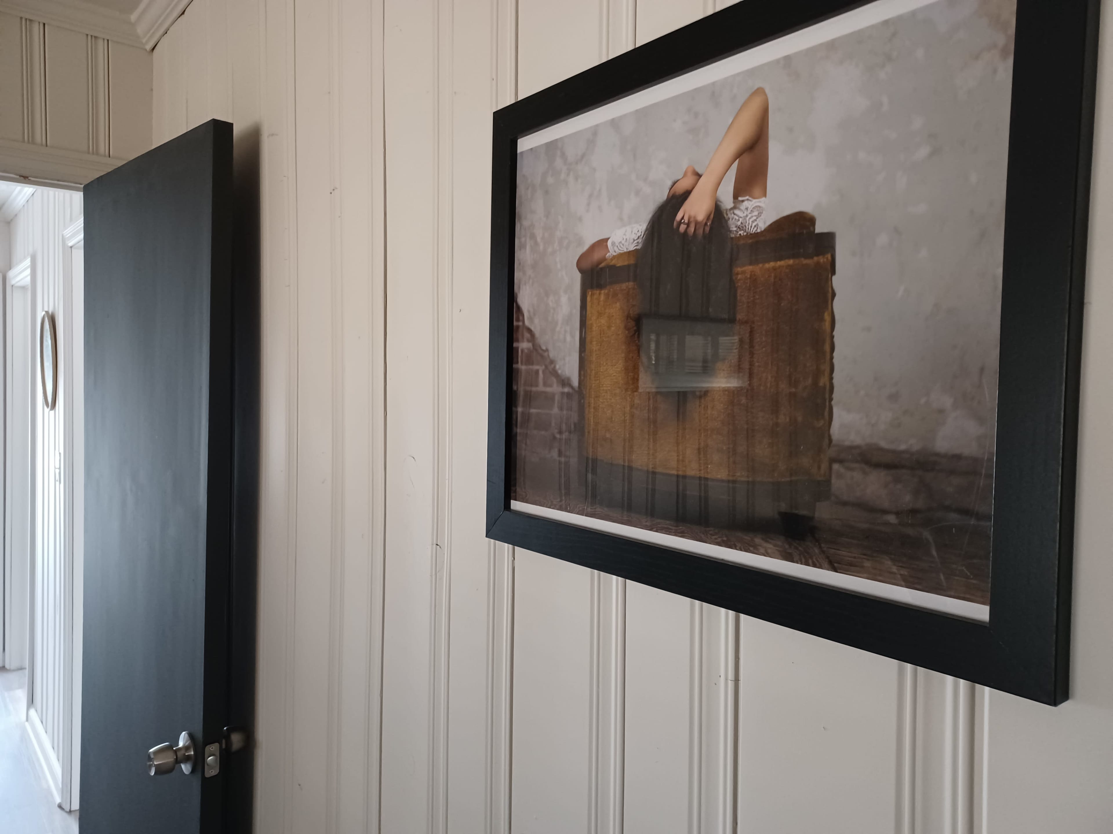 A framed photograph of a person lying on a sofa is mounted on a wall in a room with white paneling and a partially open door.