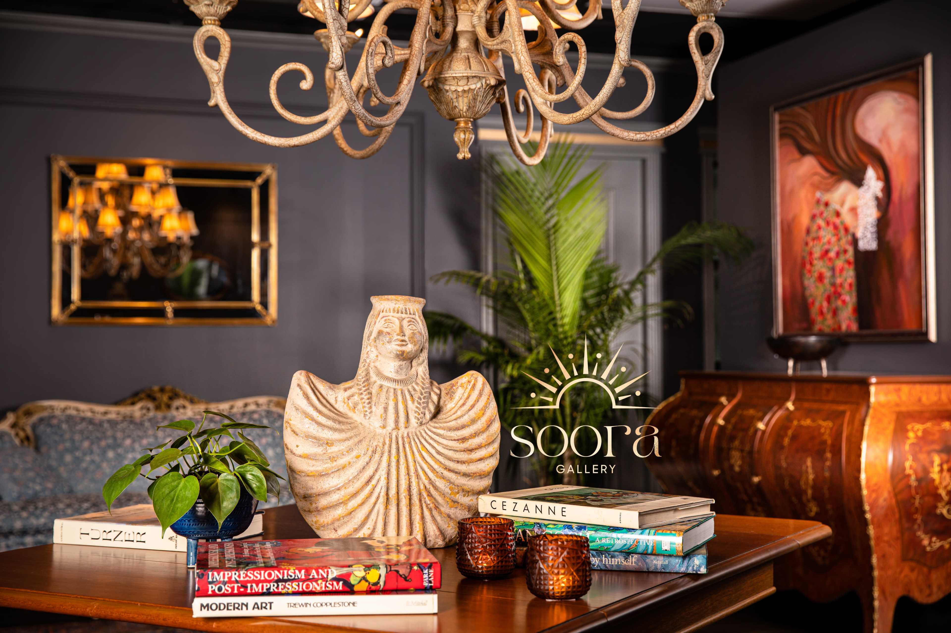 The image features a decorative interior scene with a wooden table displaying books, a plant, and a sculptural piece, complemented by a chandelier and an ornate wall painting in the background.