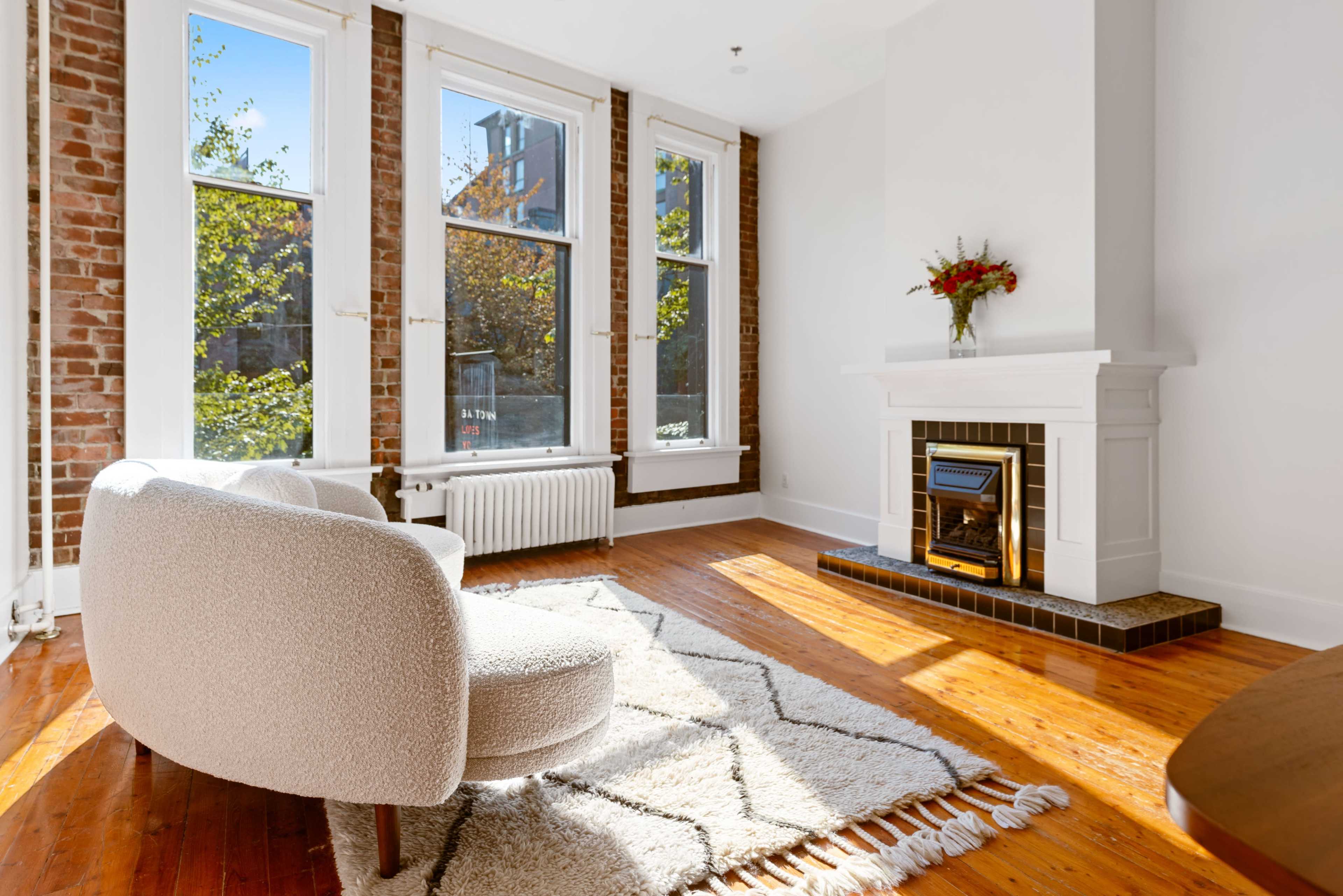 A cozy living room features a light-colored lounge chair, a patterned rug, and a fireplace with a decorative mantel beneath large windows.