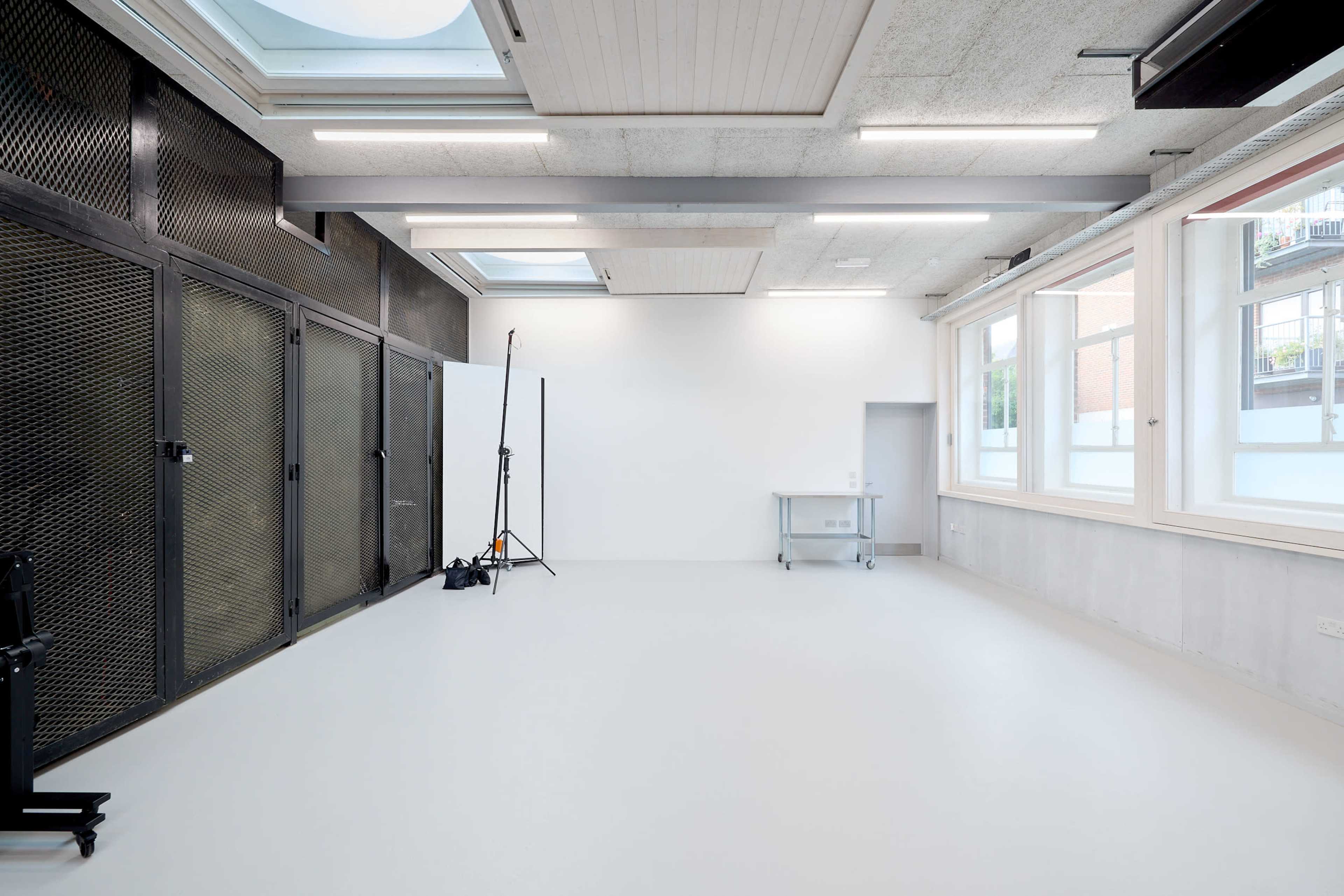 The image features a spacious, well-lit photography studio with a gray floor, high ceilings, and large windows, equipped with a backdrop stand and a movable cart.