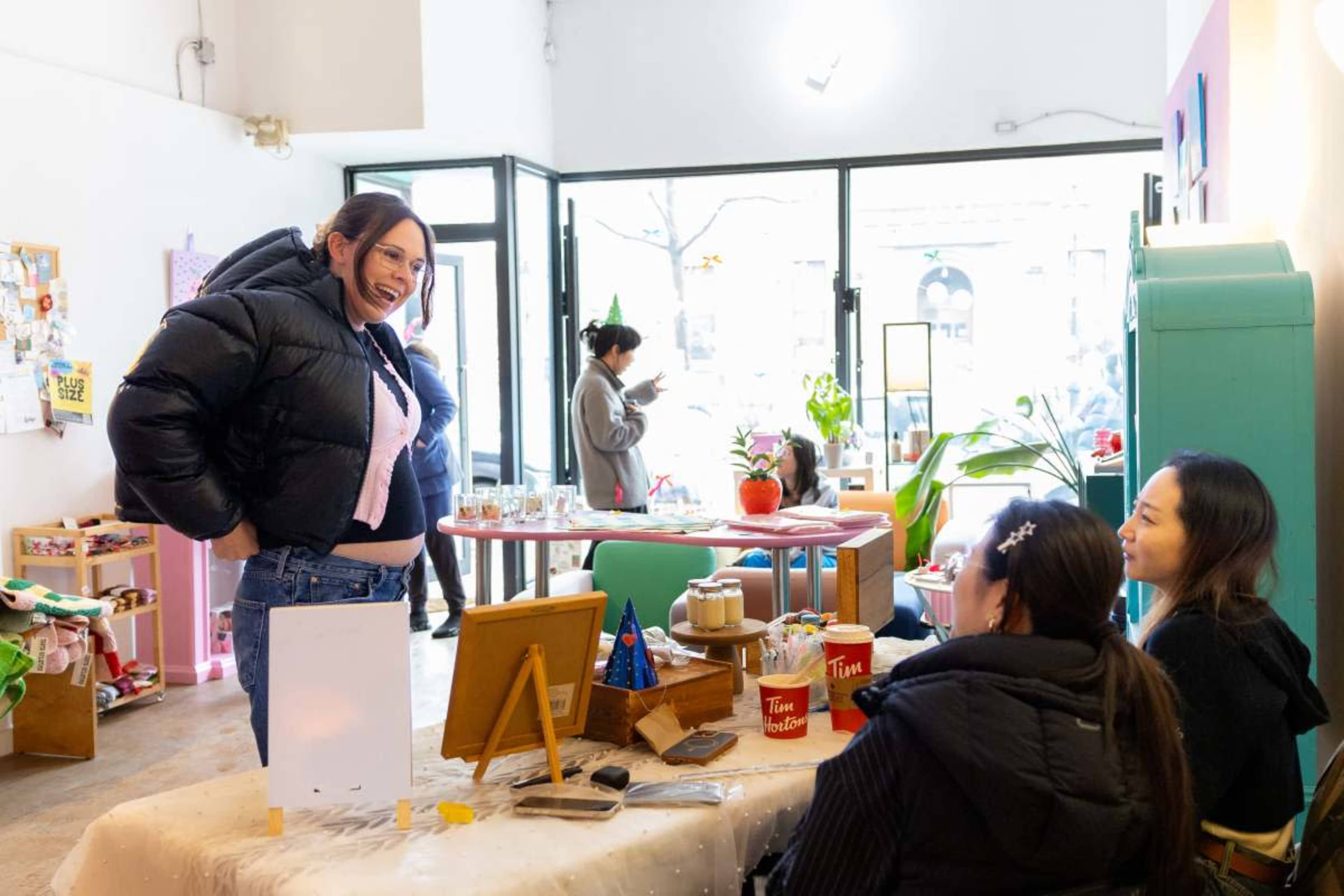 A person in a puffy jacket smiles while posing near a table with two seated individuals in a cozy cafe or store setting.