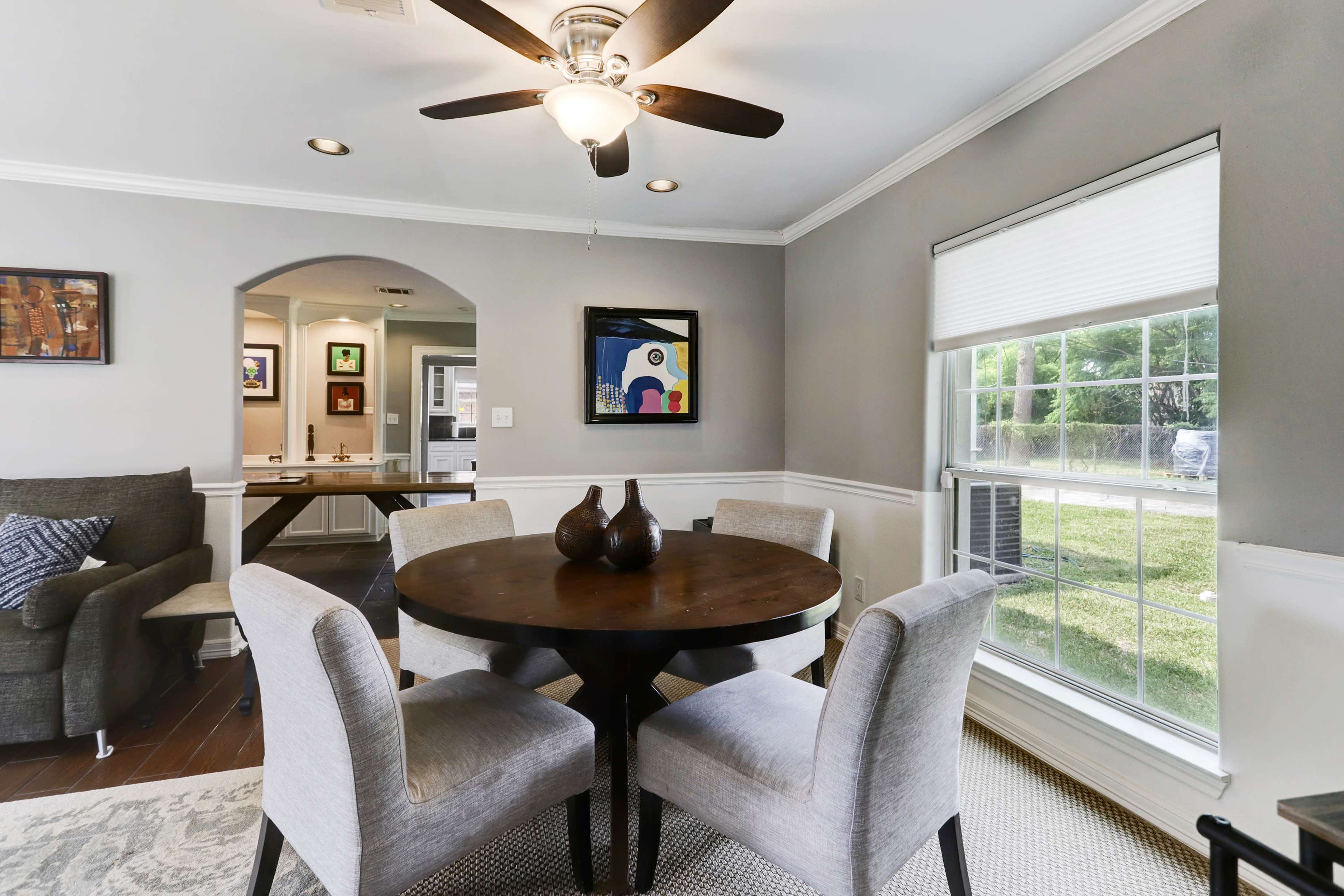 A round dining table is set with four upholstered chairs in a well-lit room featuring a ceiling fan and a window with a view of the outside.