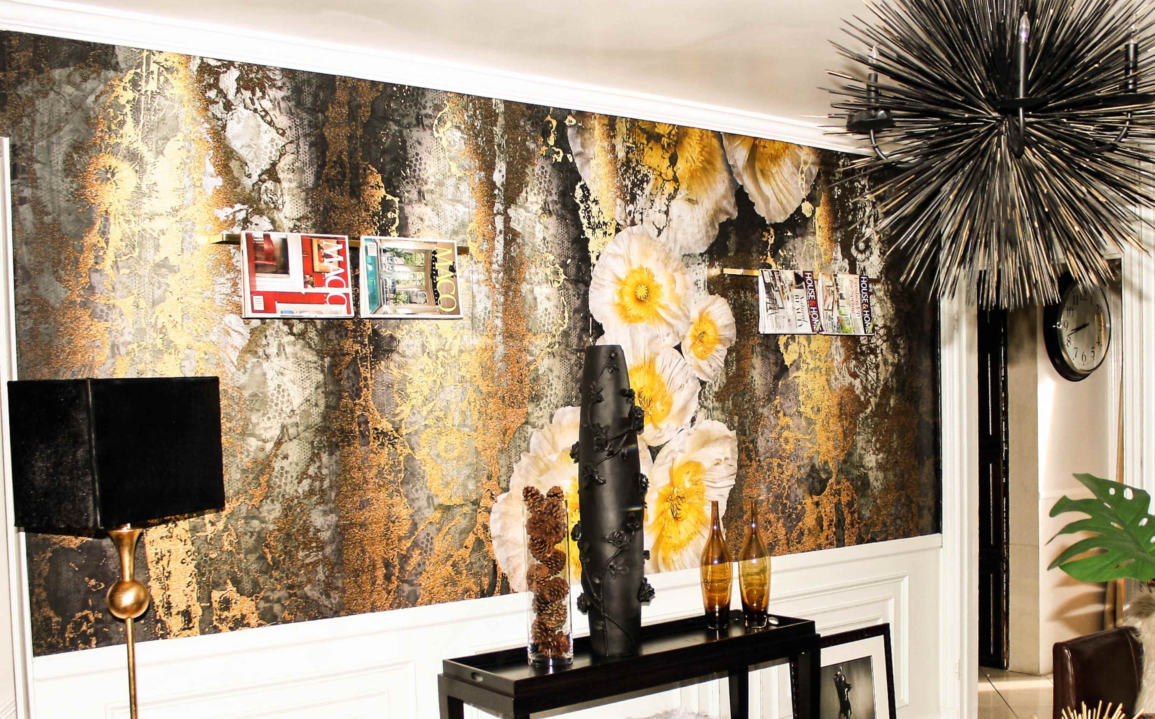 The image shows a living space with a textured wall featuring gold and dark floral patterns, complemented by a modern chandelier, a black lamp, and decorative items on a console table.
