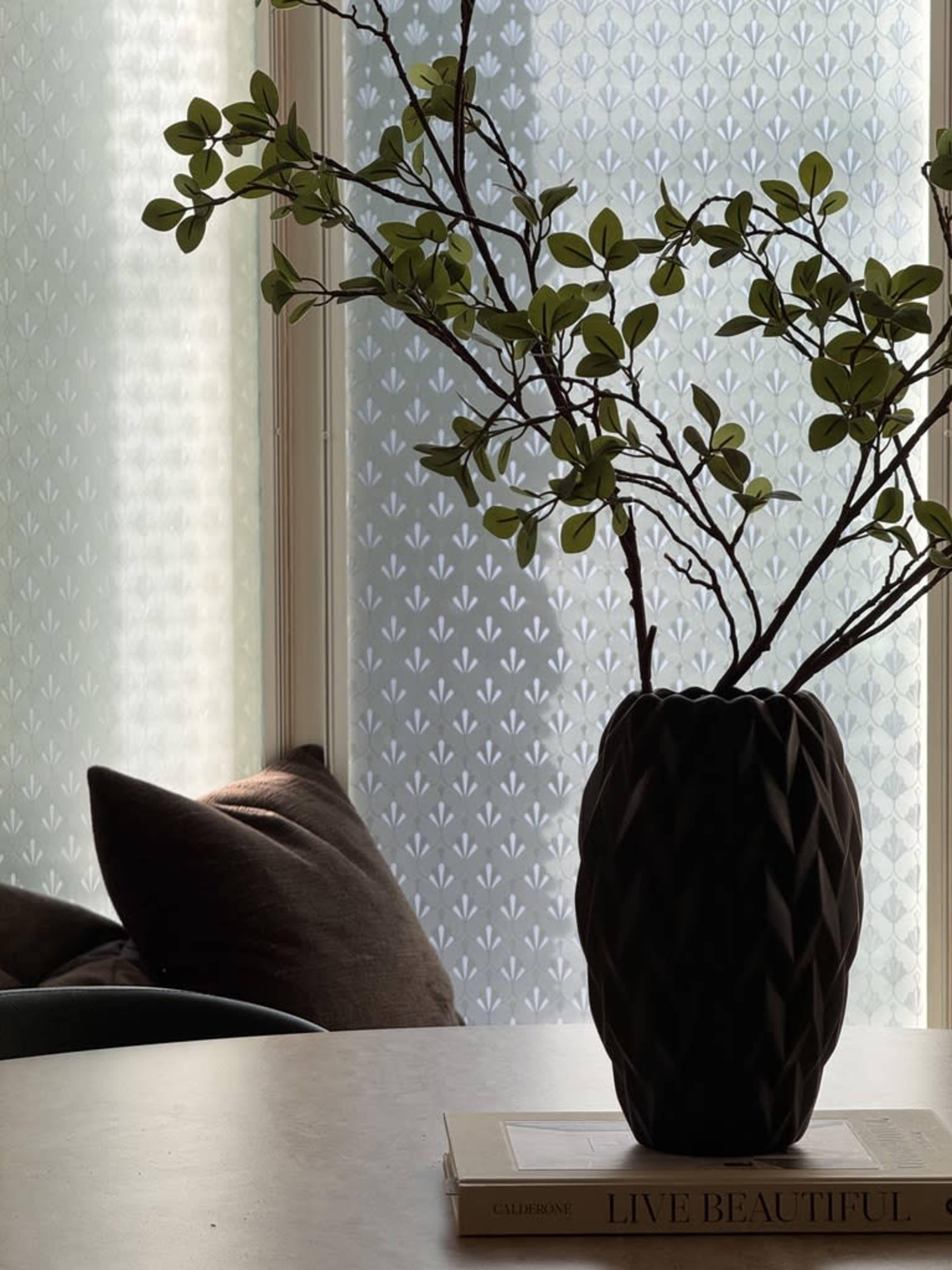 A decorative vase with branches is placed on a table next to pillows and a patterned window.