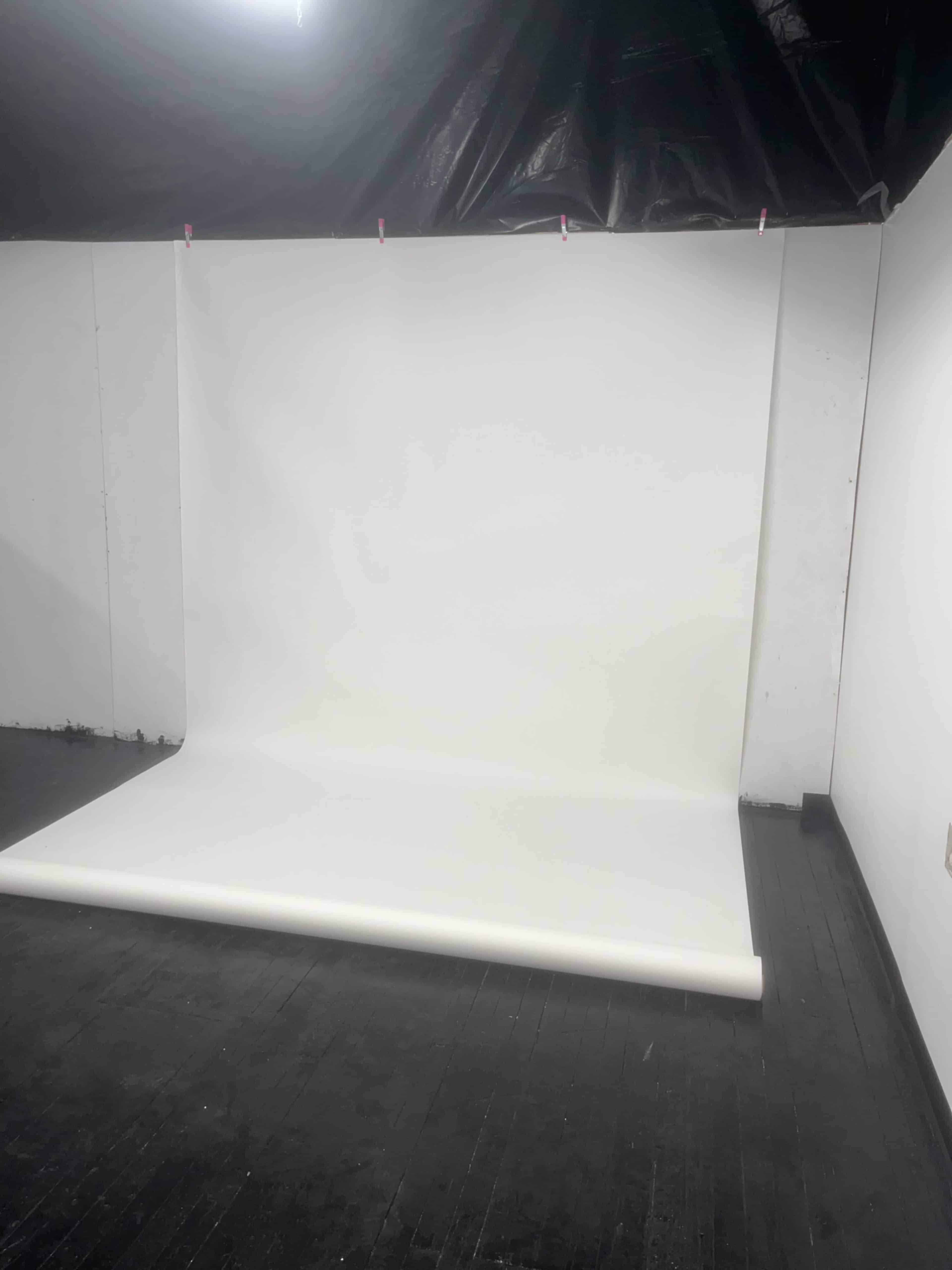 The image shows a plain white backdrop set up in a room with black flooring and partially blacked-out ceiling.