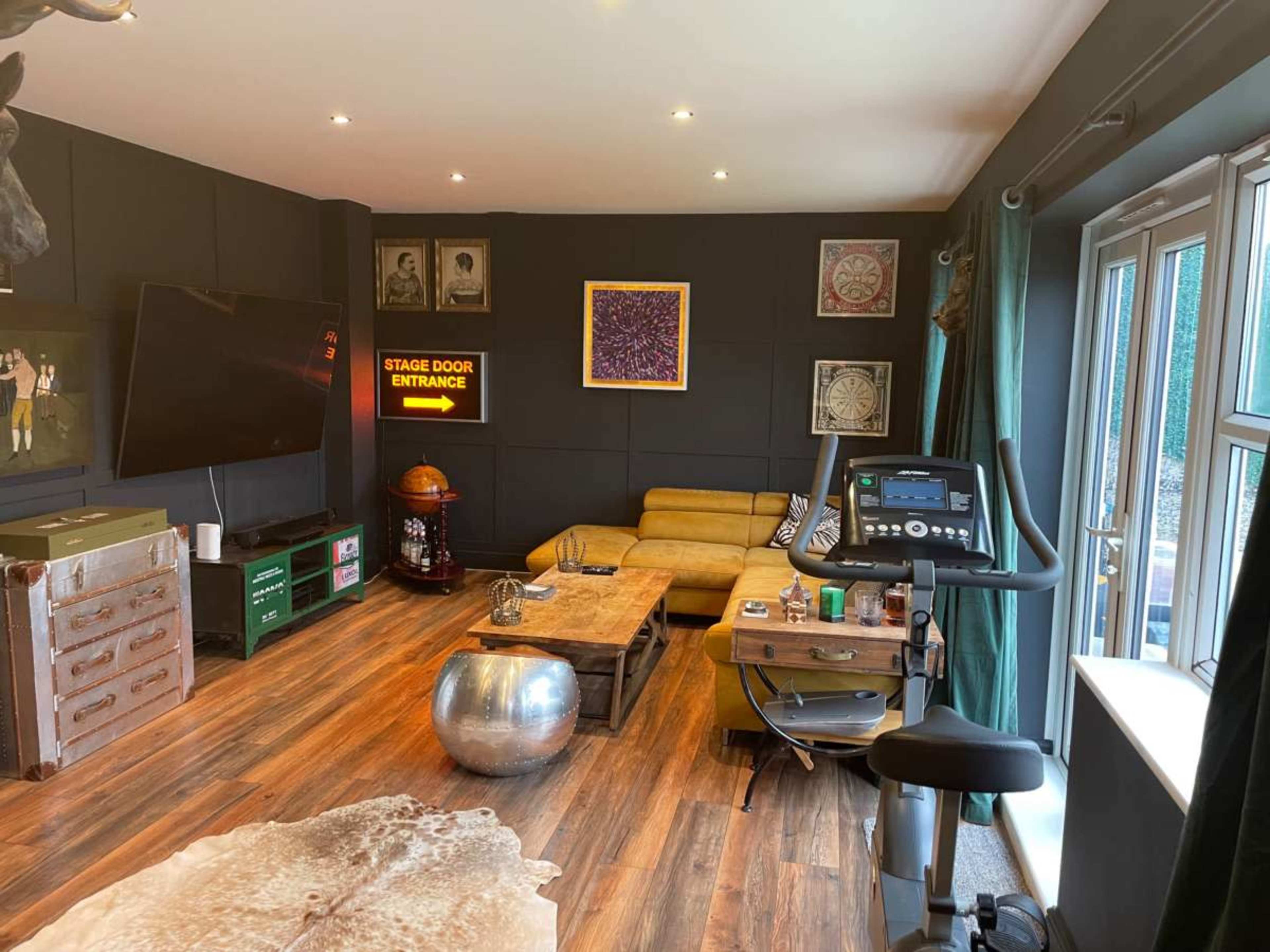 The image shows a modern living space featuring a yellow sofa, an exercise bike, a television, and various decorative items on the walls and tables.