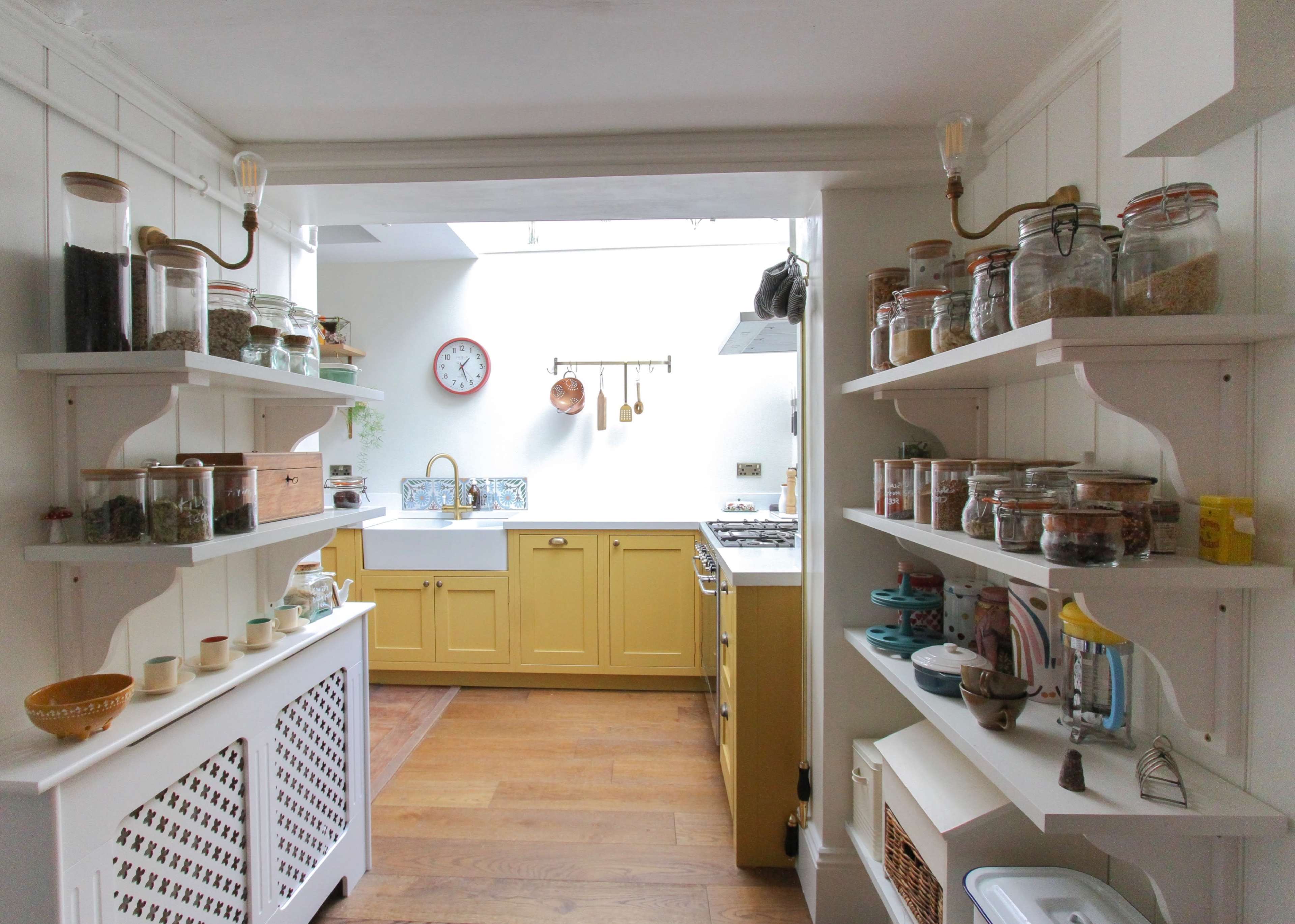 A bright kitchen features yellow cabinets, open shelves with jars of various contents, and a view through an archway into the cooking area.