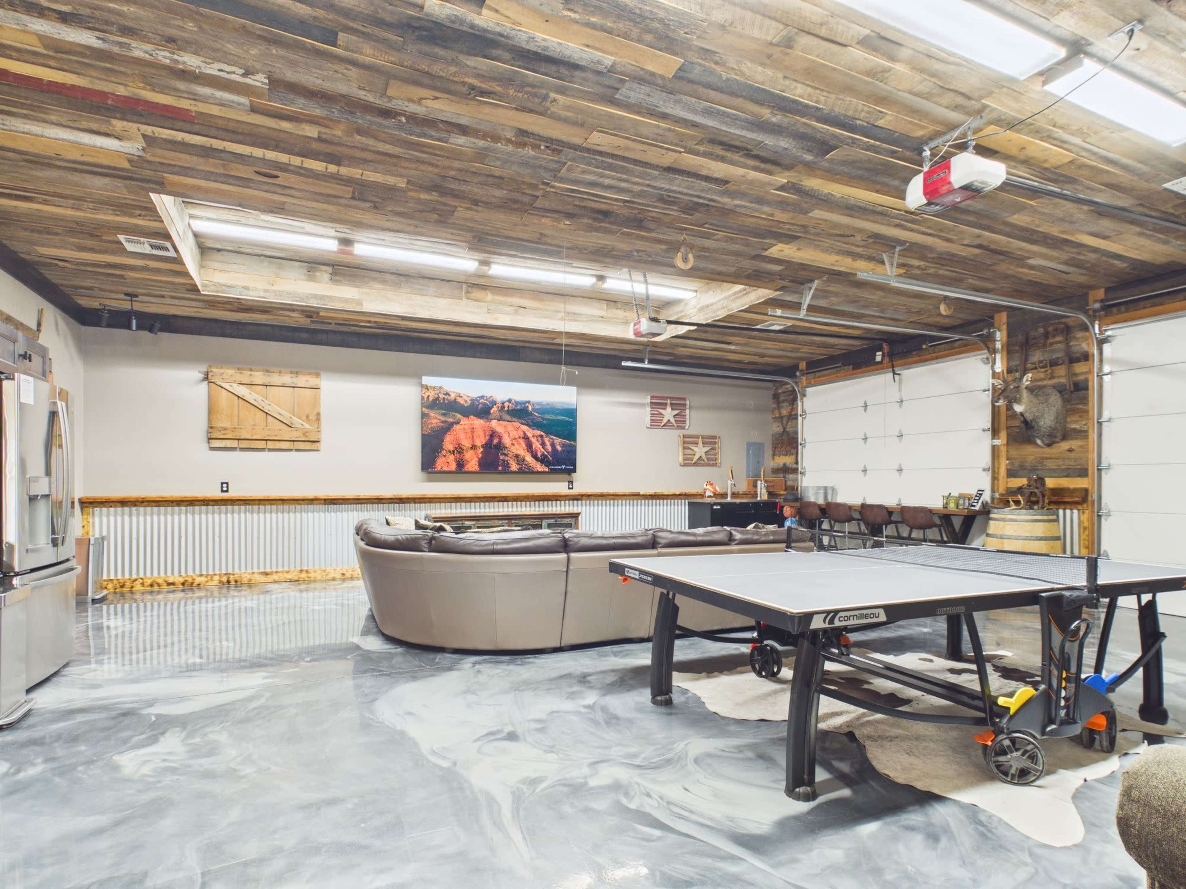 The image shows a spacious garage with a ping pong table, a large sofa, and a wooden ceiling, complemented by wall decorations and a garage door at one end.