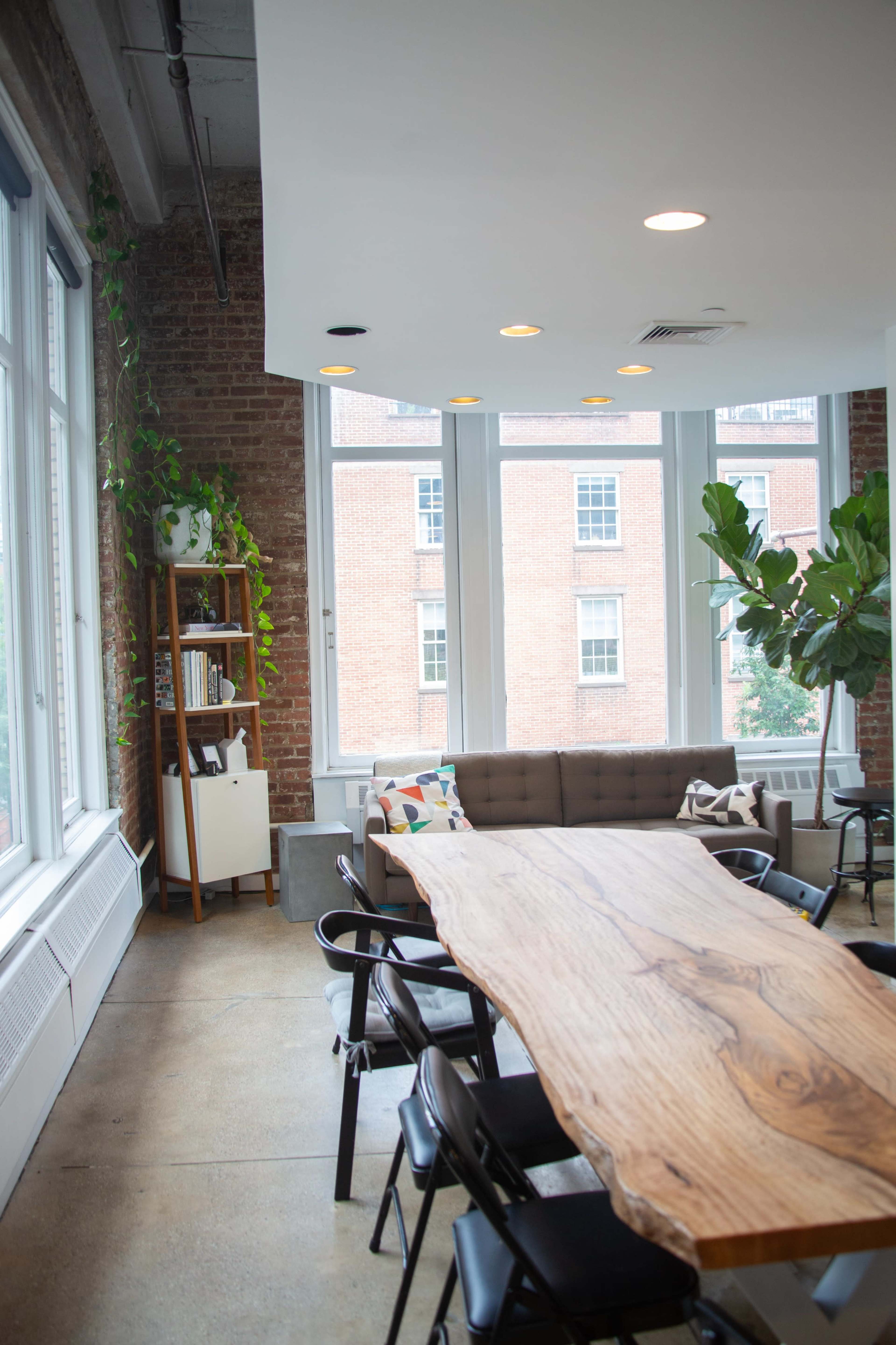 The image shows a modern meeting space featuring a large wooden table surrounded by black chairs, a couch, and large windows with views of brick buildings outside.