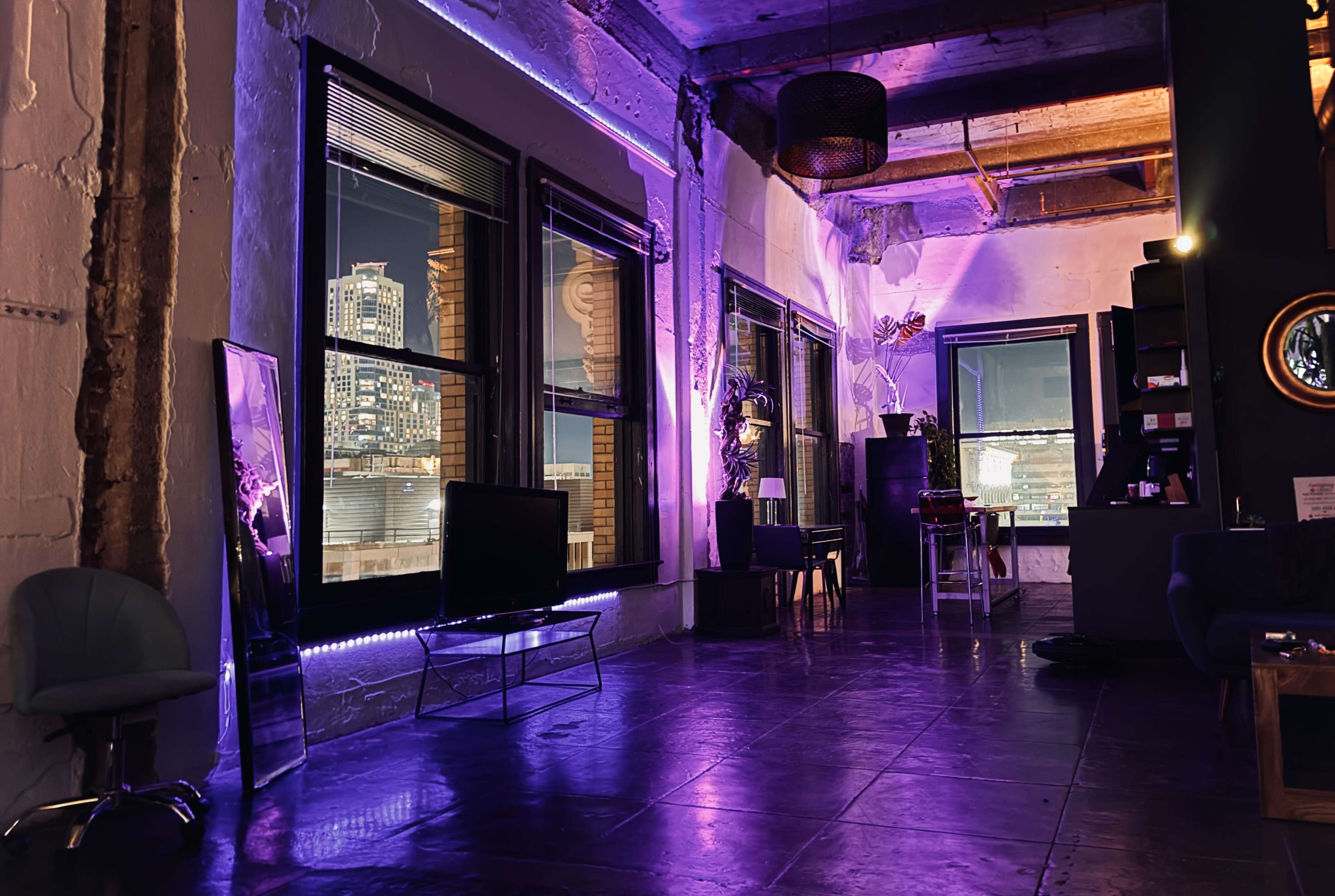 A modern interior space features large windows with city views, illuminated by purple LED lights and minimalist furniture.