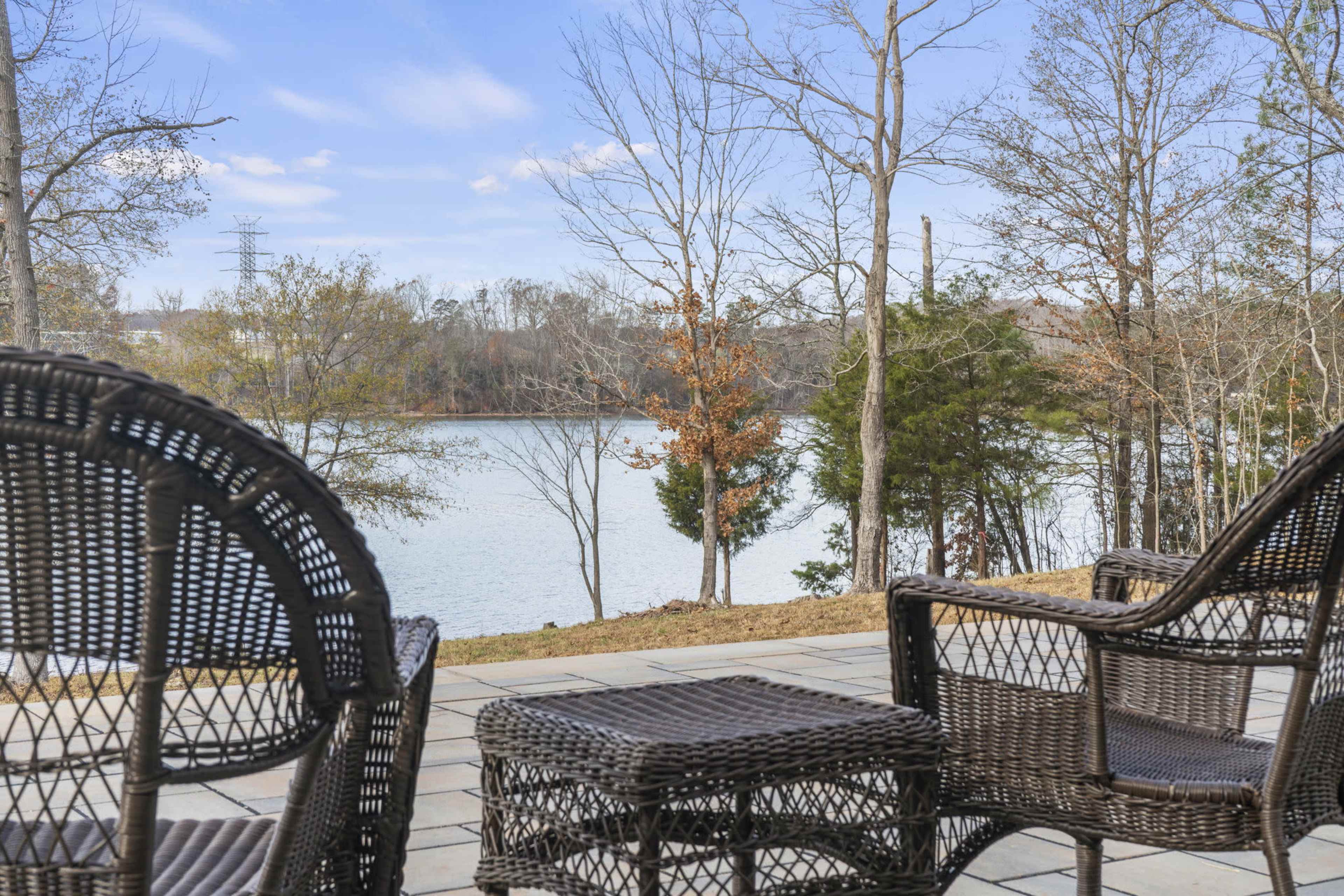 A patio area with wicker chairs overlooks a calm lake surrounded by trees.