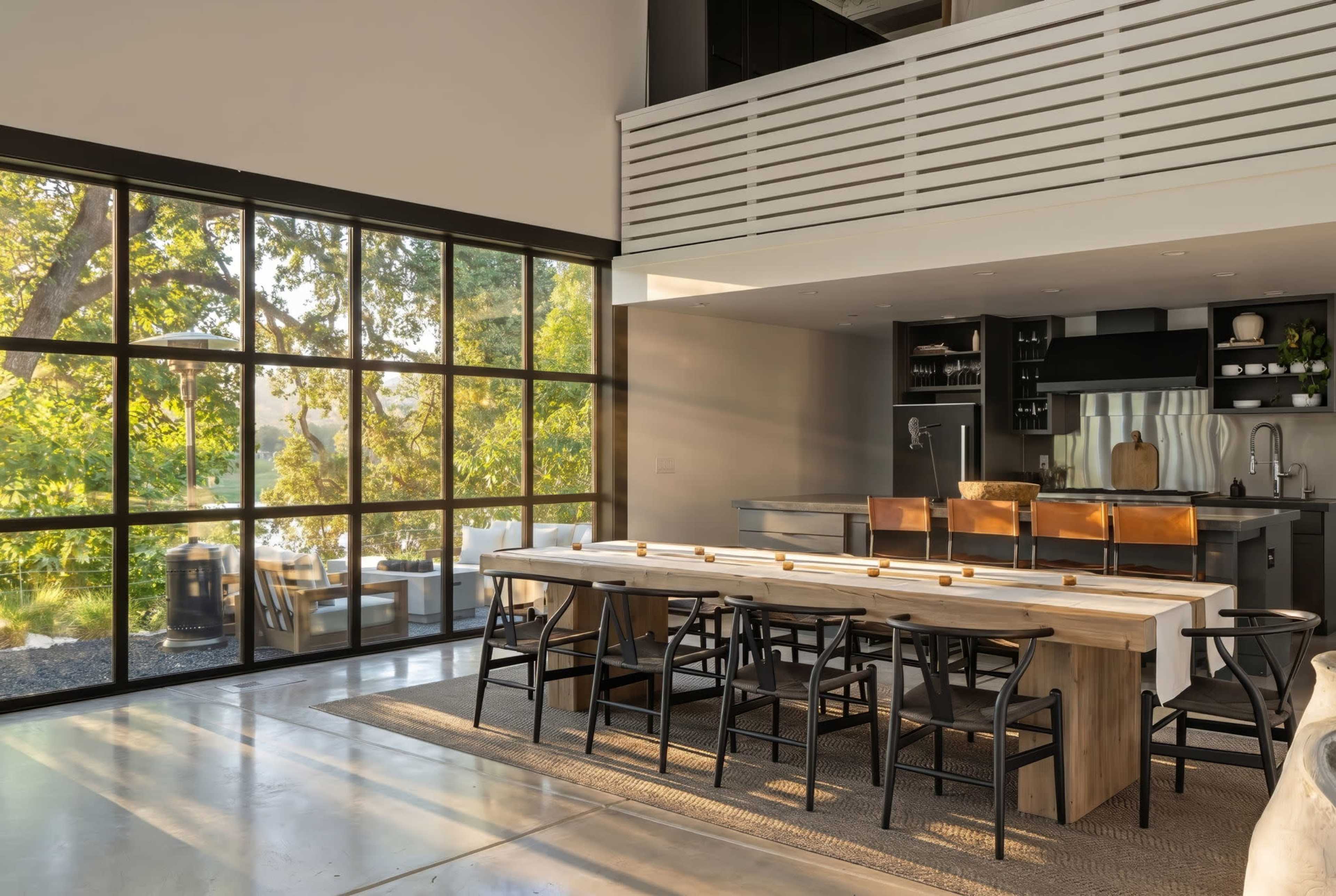 The image shows a modern kitchen and dining area with a large wooden table, black chairs, and large windows letting in natural light.