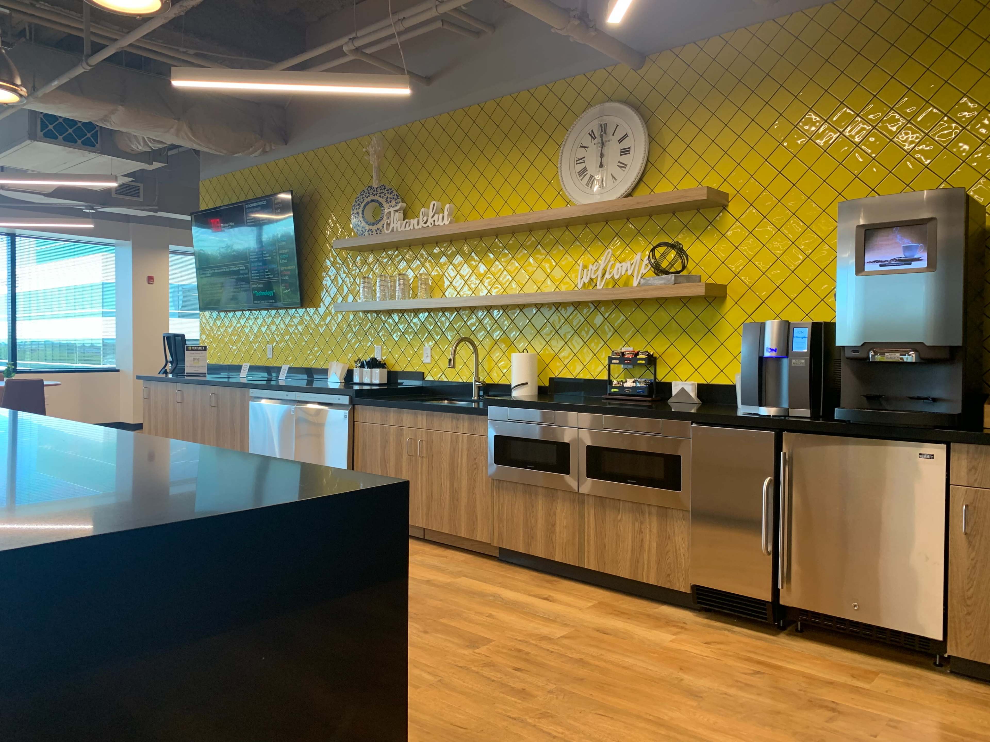 The image shows a modern kitchen area with yellow tiled walls, stainless steel appliances, and wooden cabinetry.