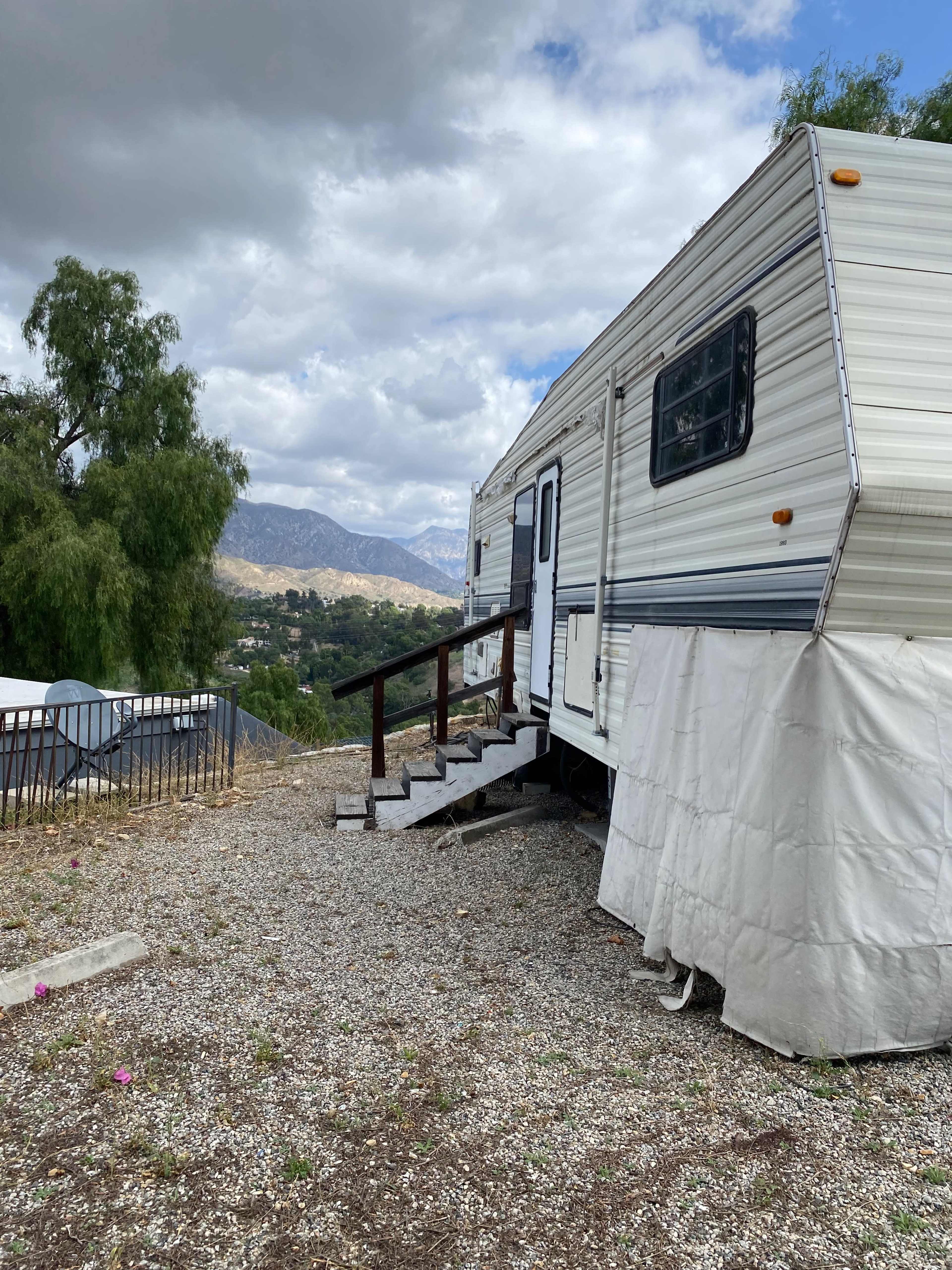 A mobile home is positioned on a gravel lot with wooden steps leading up to its door, surrounded by trees and distant hills under a cloudy sky.