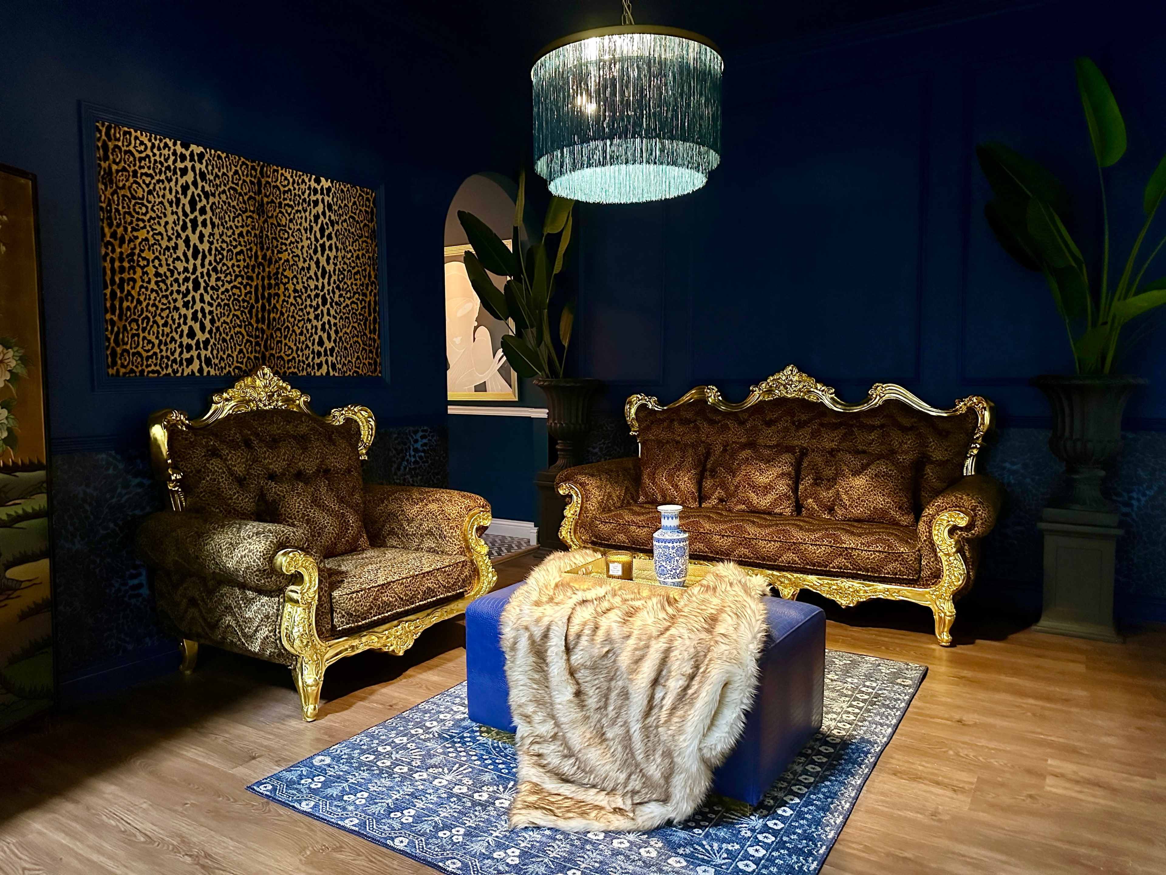 The room features two ornate gold-patterned sofas, a blue coffee table topped with a fur throw, and a large chandelier, all set against a dark blue wall.