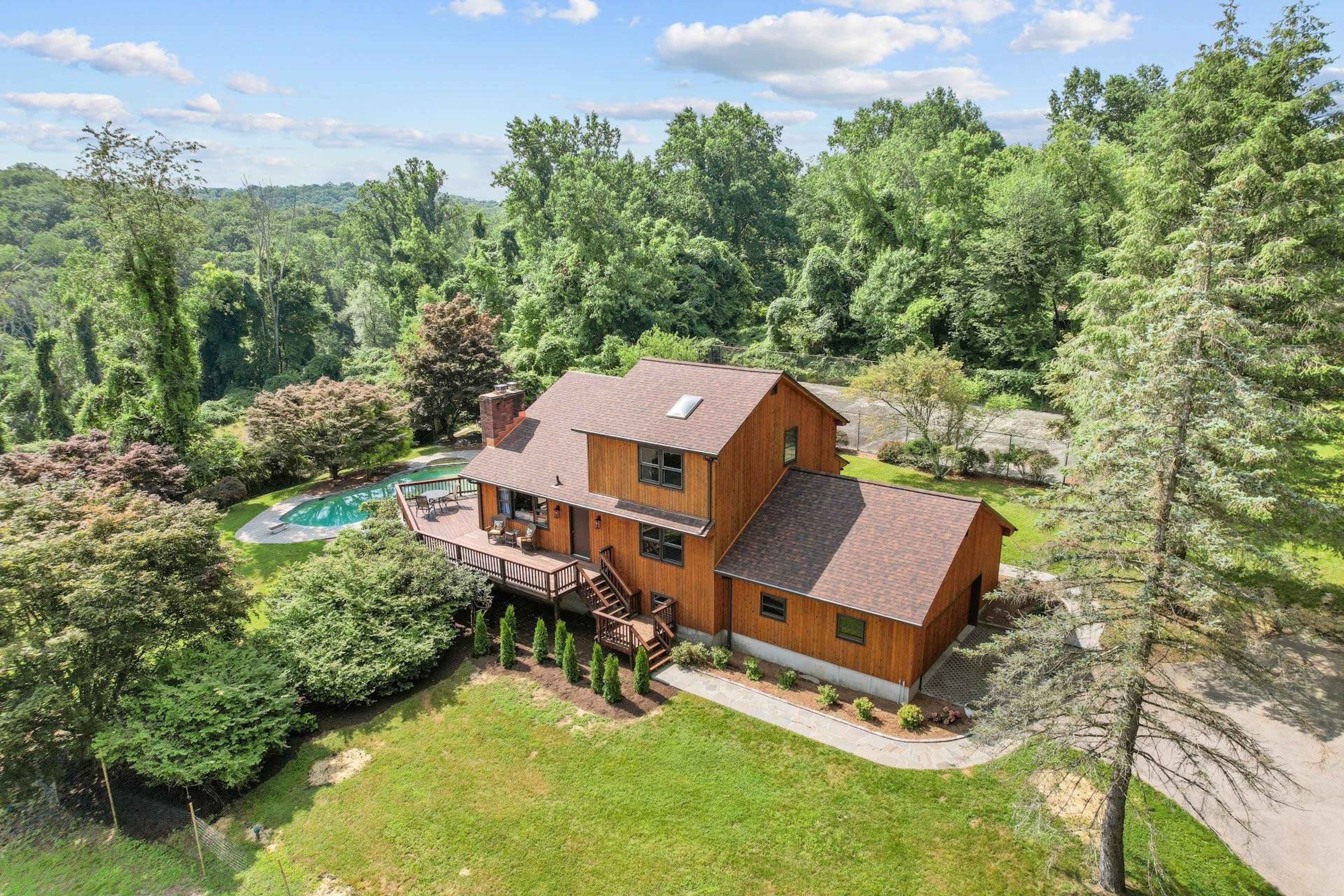 A two-story wooden house with a swimming pool surrounded by trees and landscaped gardens.