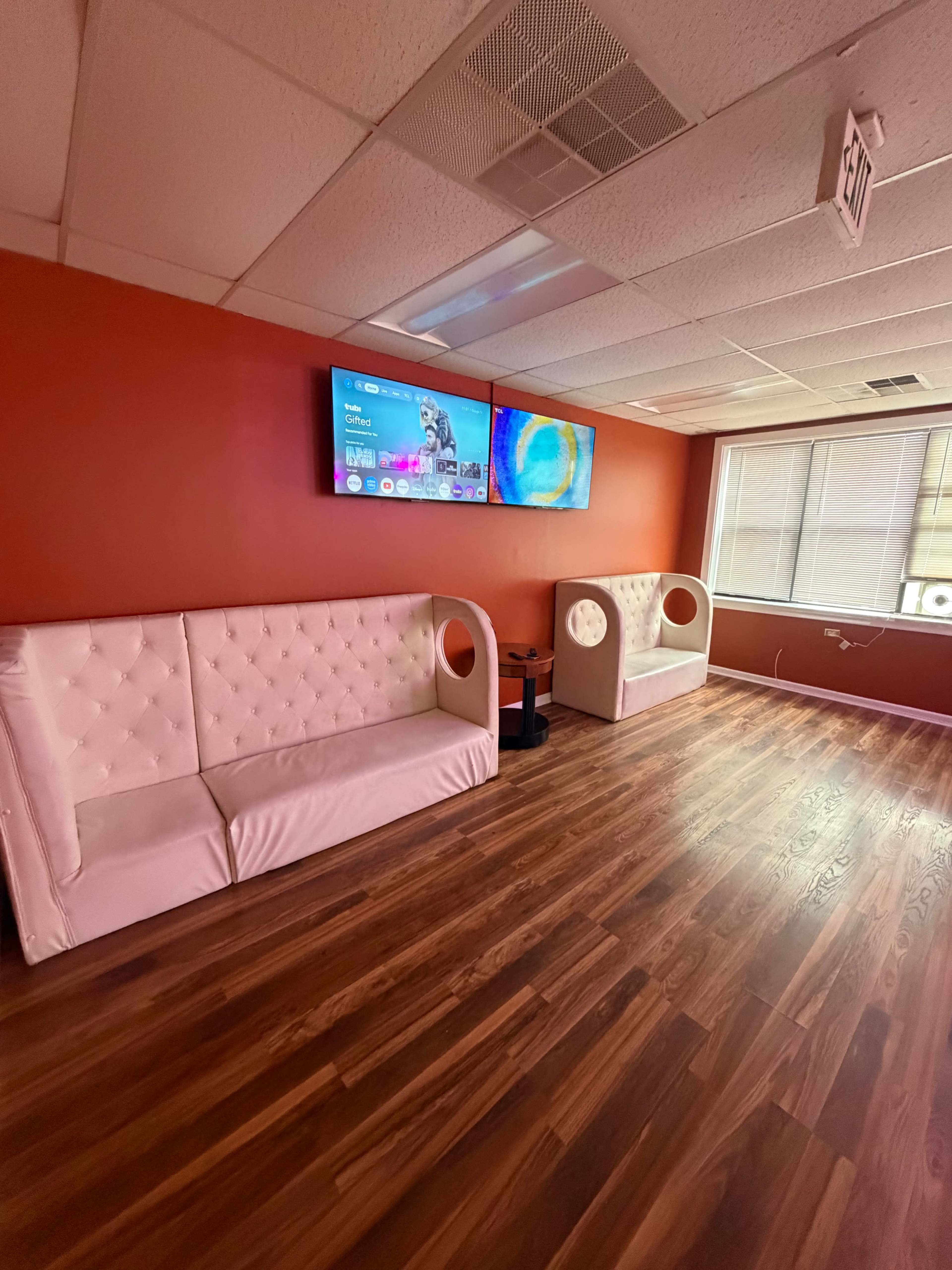 The image shows a modern lounge area with two white tufted sofas, brown wooden flooring, and two wall-mounted televisions against a red wall.