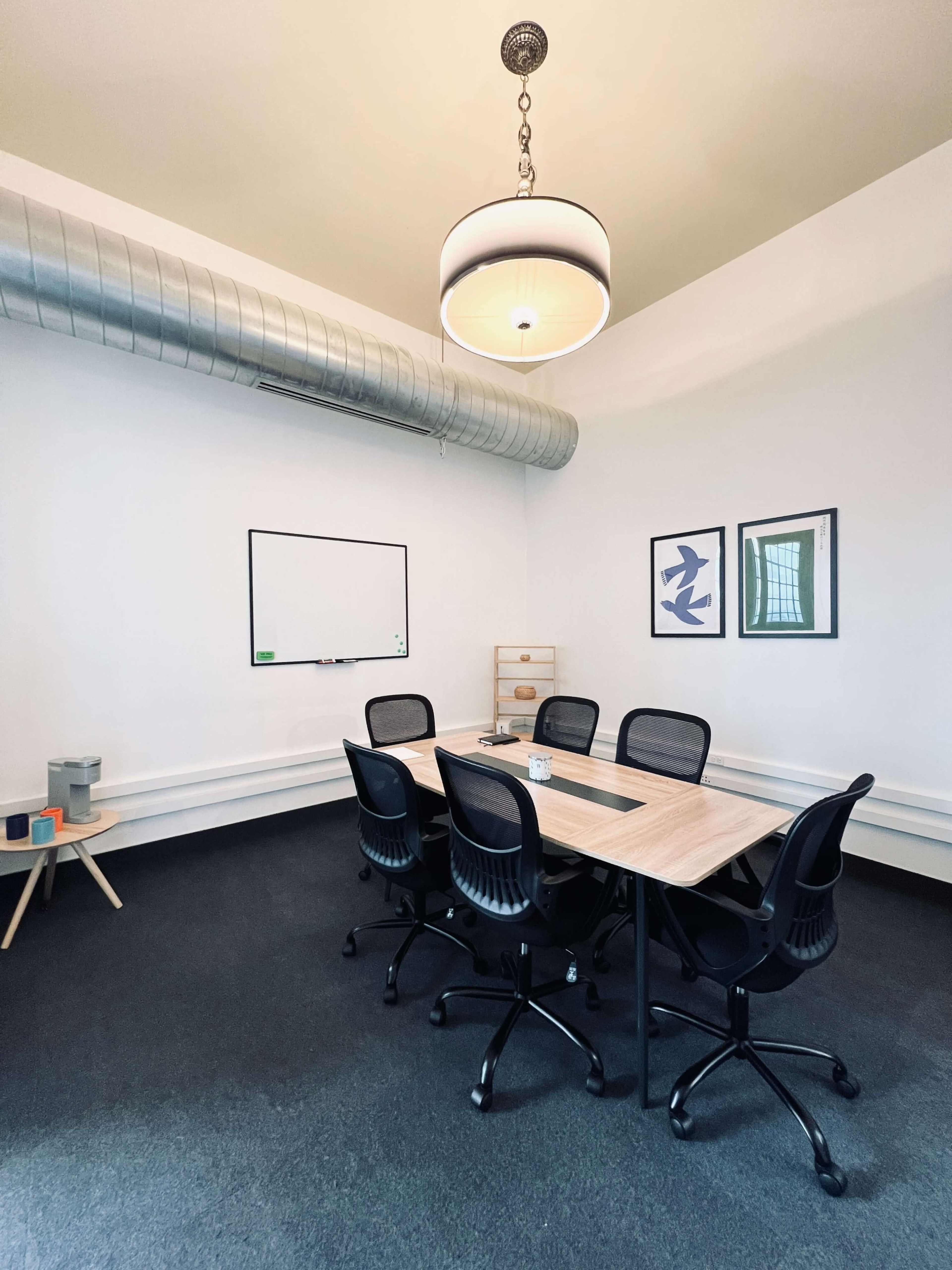 A meeting room features a rectangular table surrounded by six black chairs, with a whiteboard and artwork on the walls.