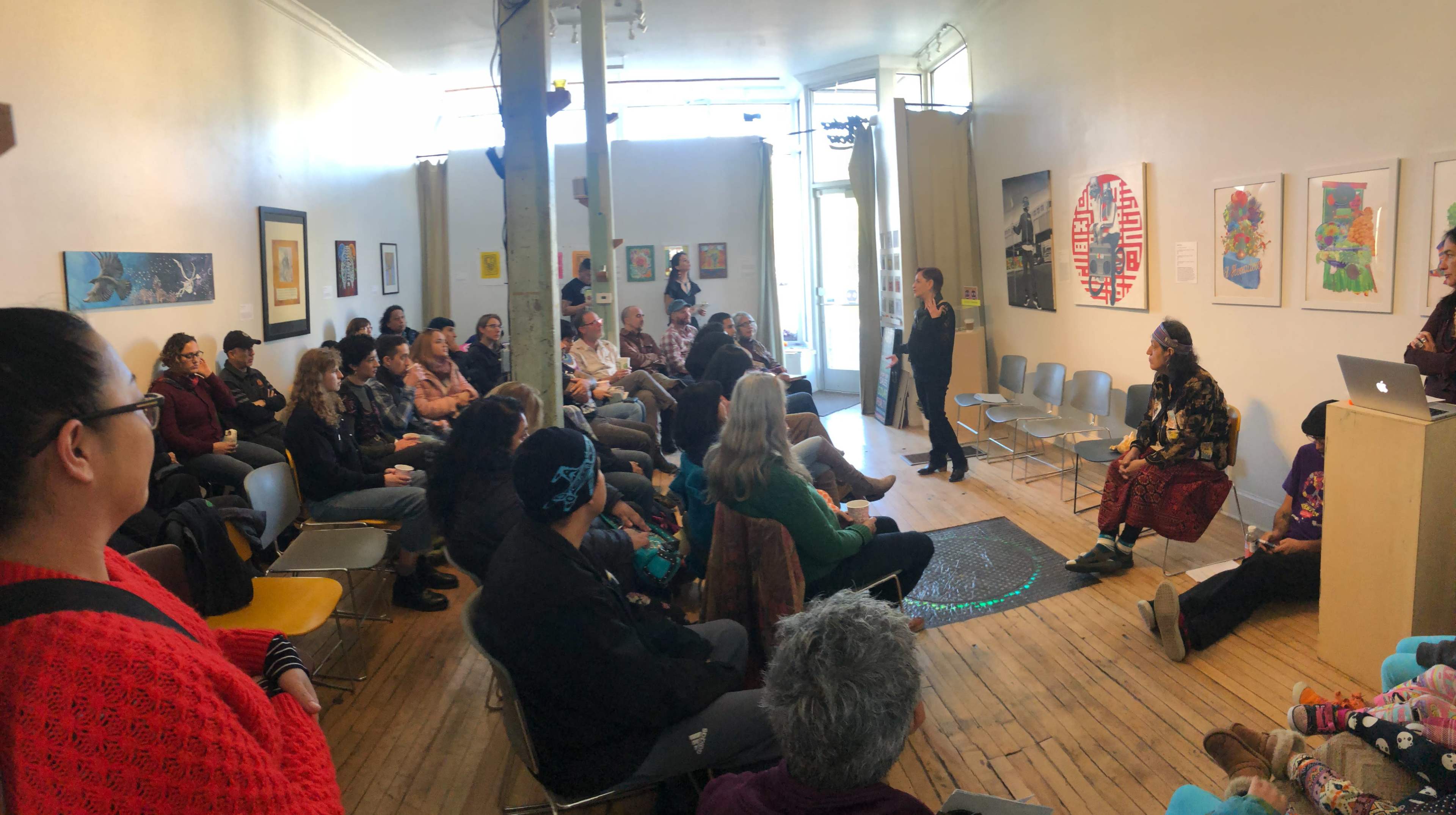 A speaker presents to an audience seated in a well-lit room with art on the walls.