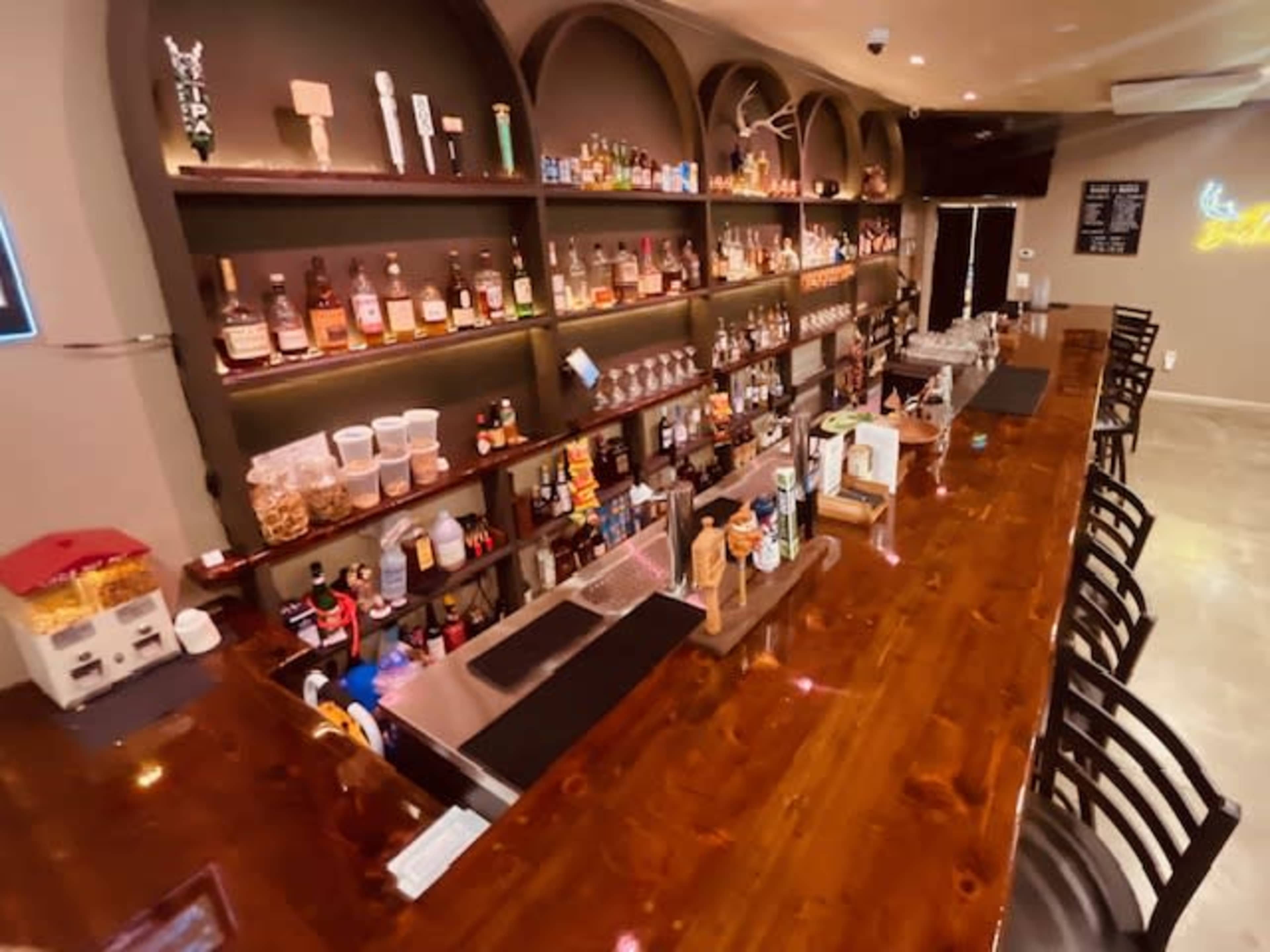 The image shows a well-stocked bar with an assortment of bottles on shelves behind a polished wooden countertop and black bar stools arranged in front.