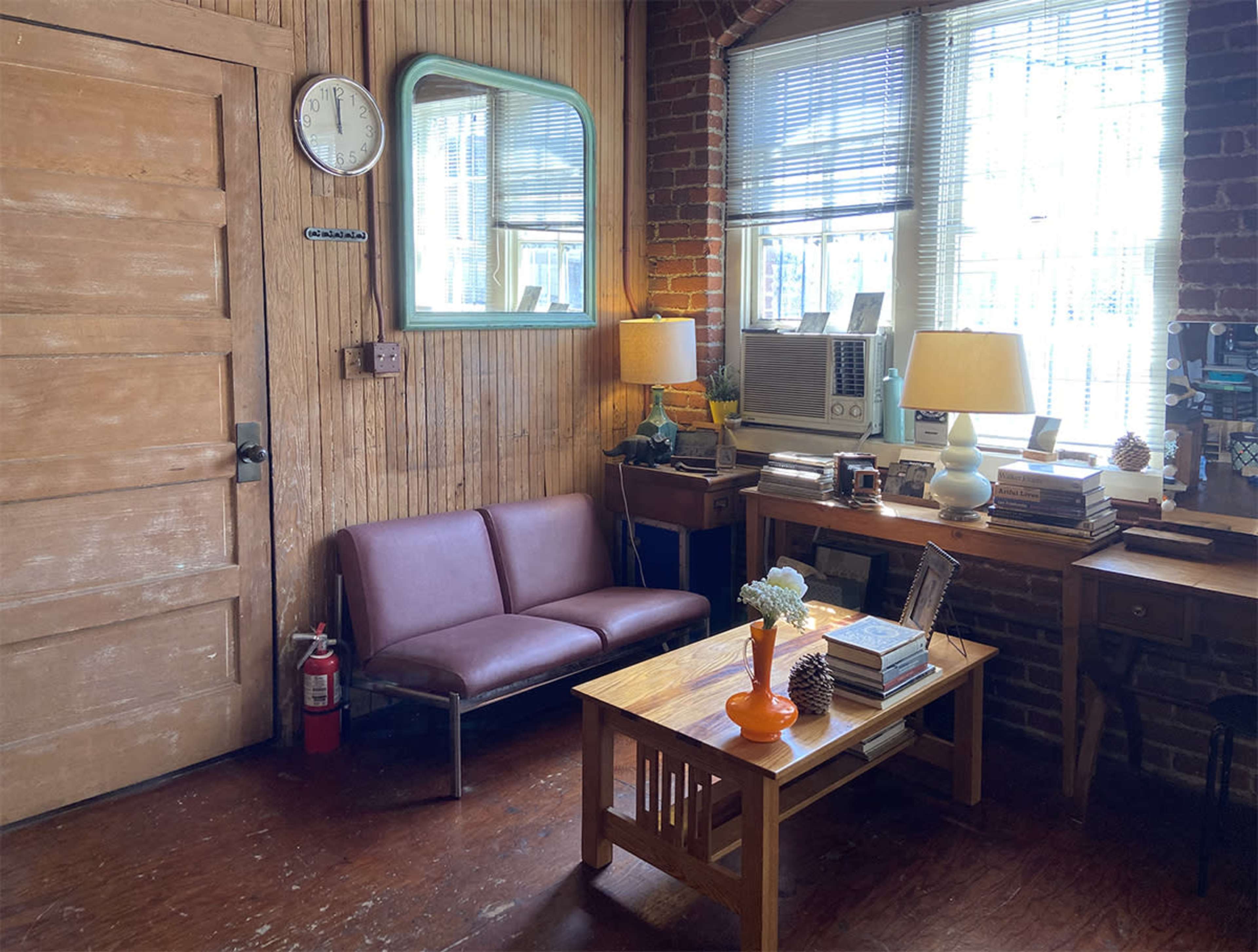 A small, cozy room with a brown couch, a wooden coffee table, and two desks by the window, featuring a clock, an air conditioner, and lamps.