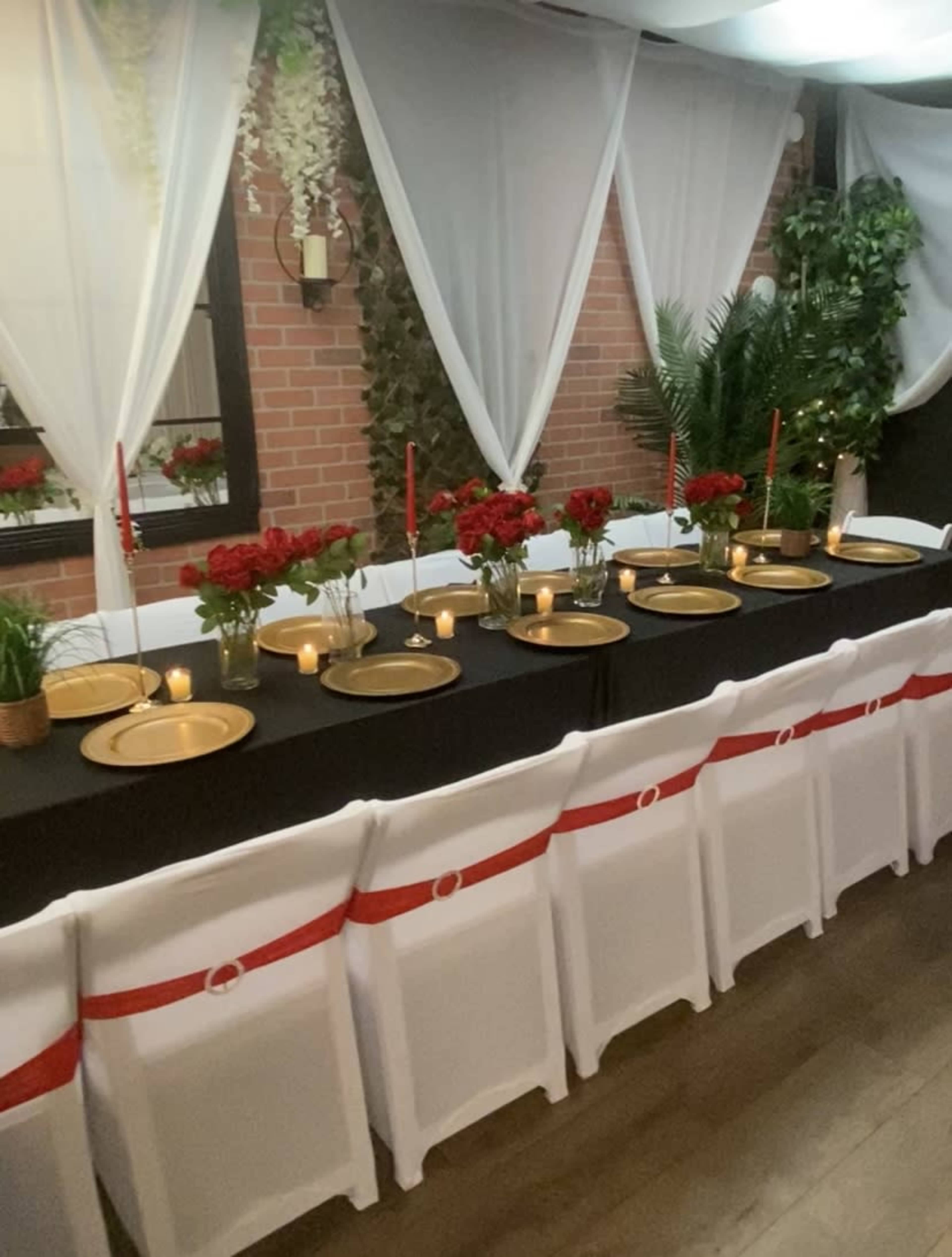 A long dining table is set in a decorated room with red roses, gold plates, candles, and white chairs adorned with red ribbons.