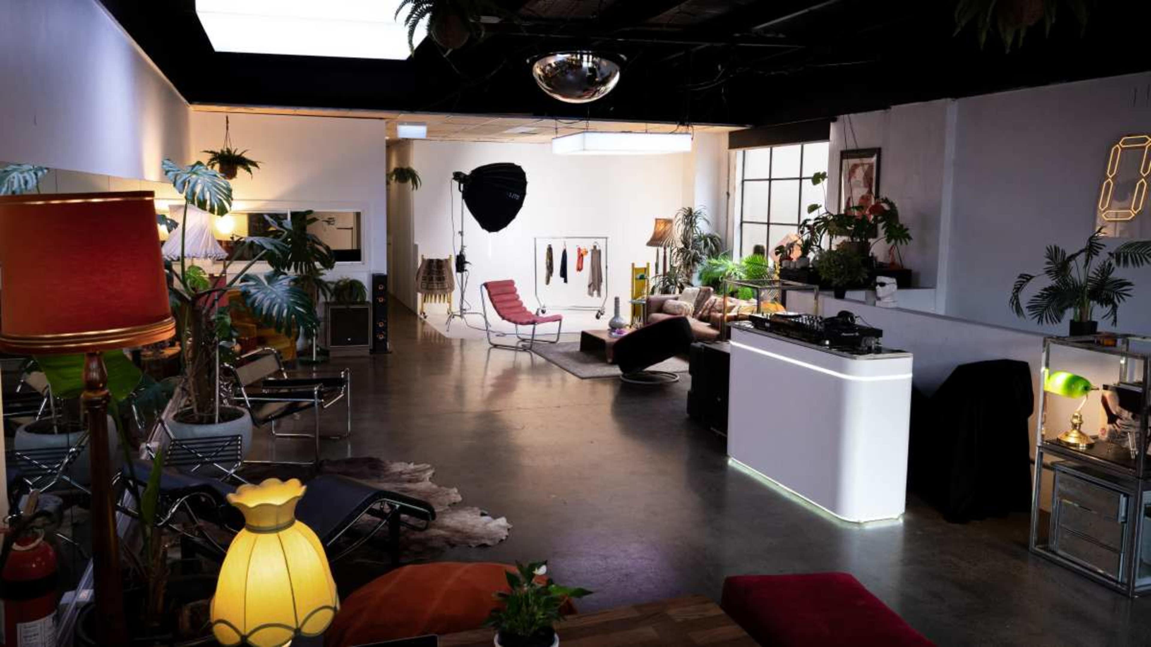 The image shows a modern indoor space featuring various plants, seating areas, a DJ setup, and photography equipment.
