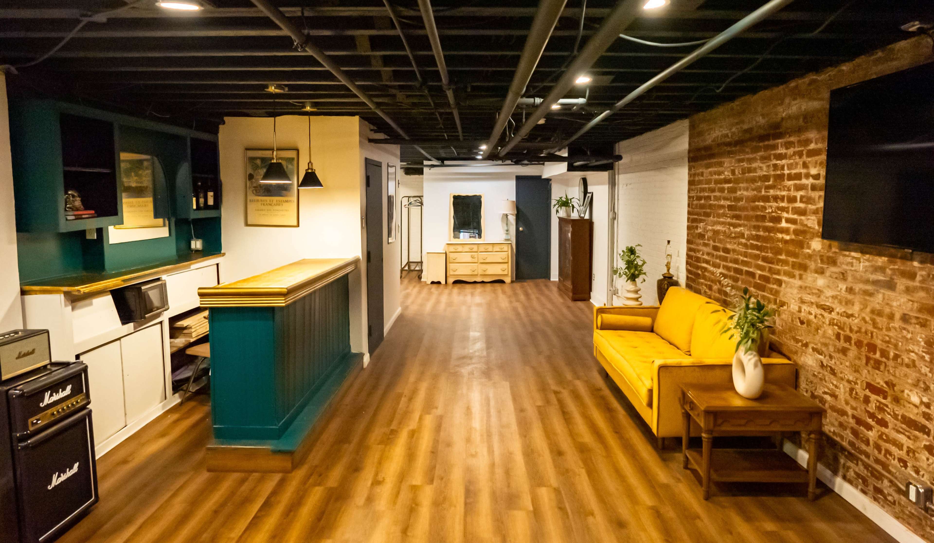A spacious interior with wooden flooring, featuring a yellow sofa, a vintage dresser, and a bar area against a teal accent wall, alongside exposed brick walls.