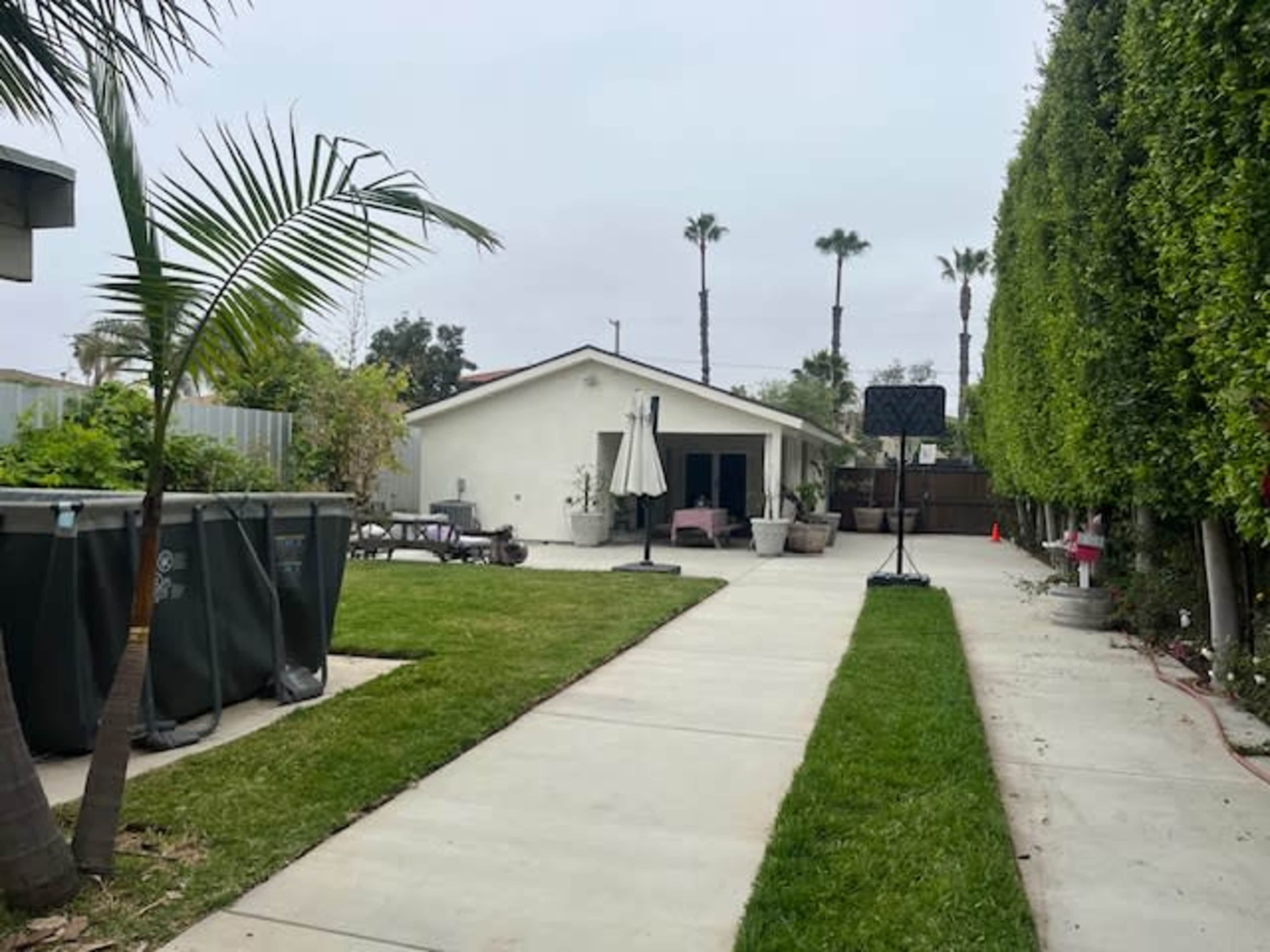 A paved walkway leads through a backyard with green grass and palm trees, flanked by potted plants and a fenced area with a basketball hoop.
