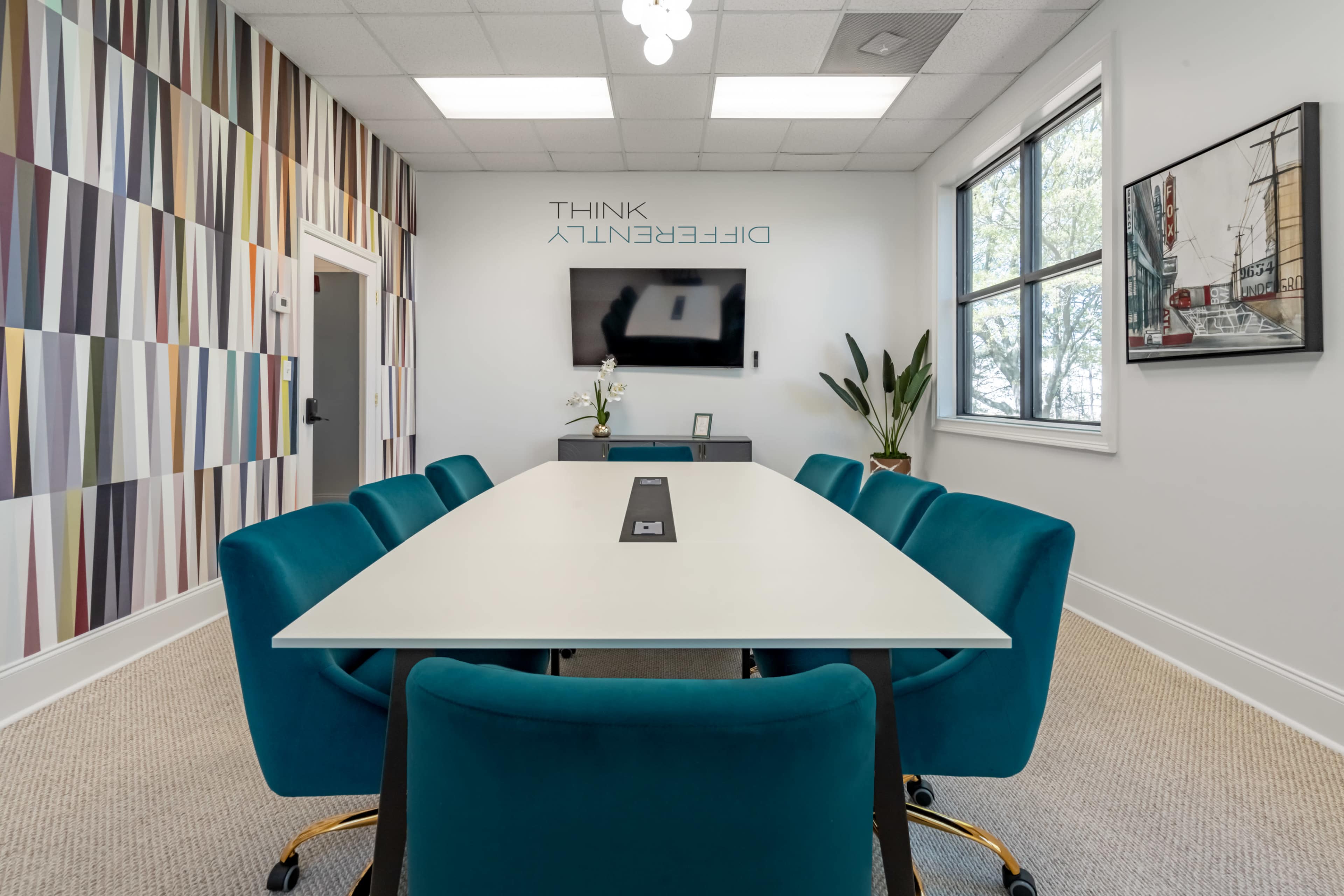 A modern conference room features a long white table surrounded by teal chairs, with a colorful patterned wall and a television mounted on the opposite side.