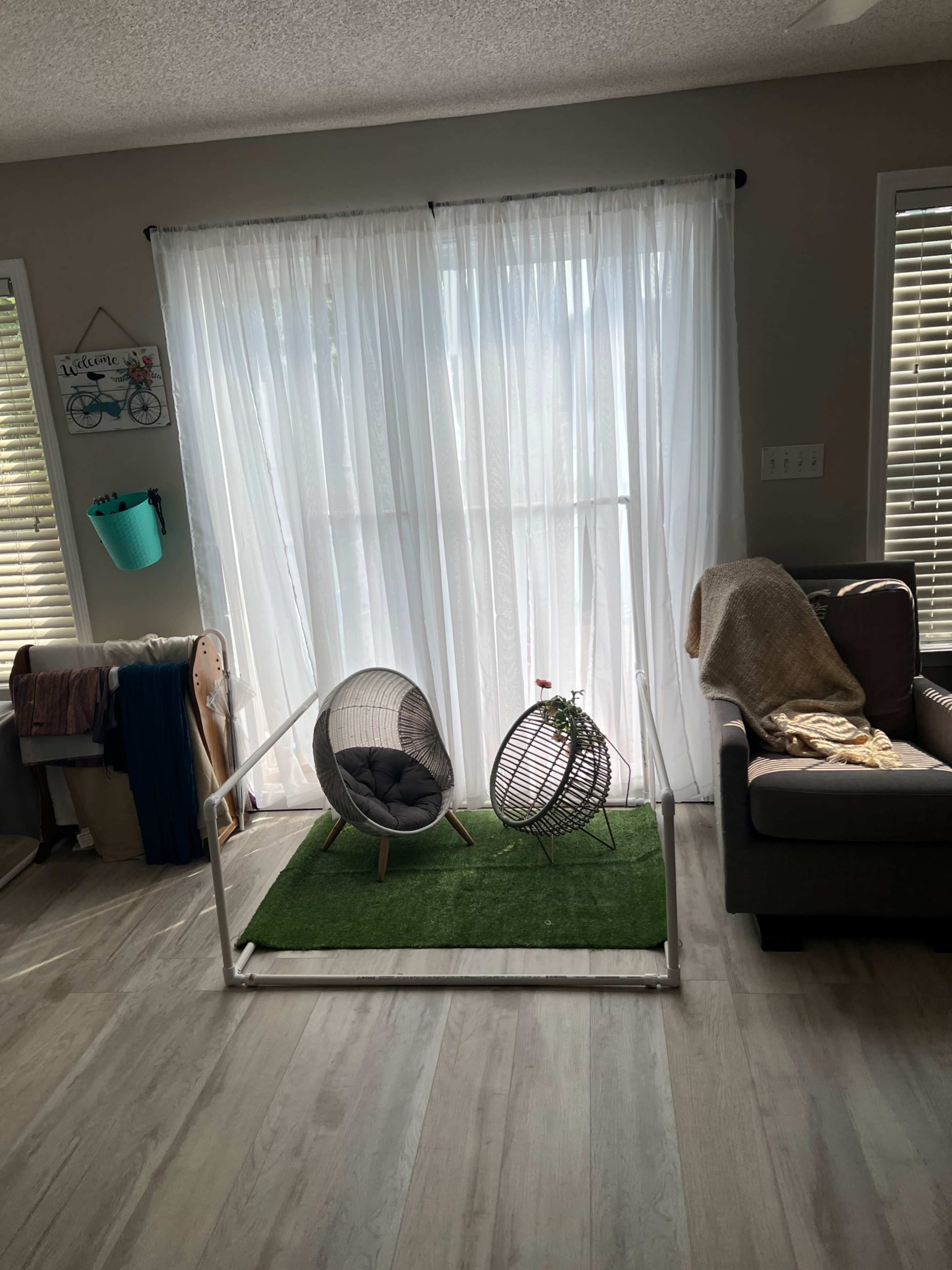 A living room features two wire chairs on a small patch of artificial grass in front of a large window with white curtains.