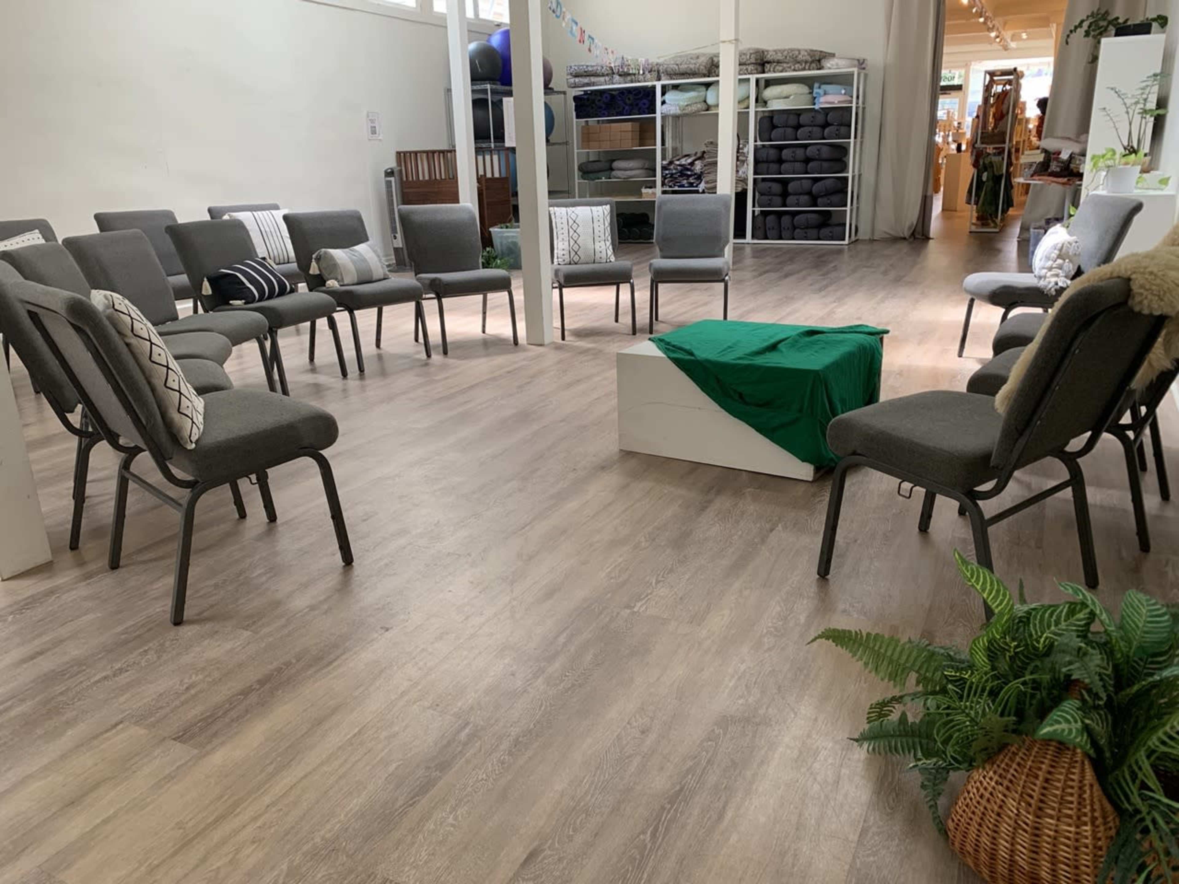 A circular arrangement of gray chairs surrounding a green cloth draped over a white platform in a well-lit room with wooden flooring.
