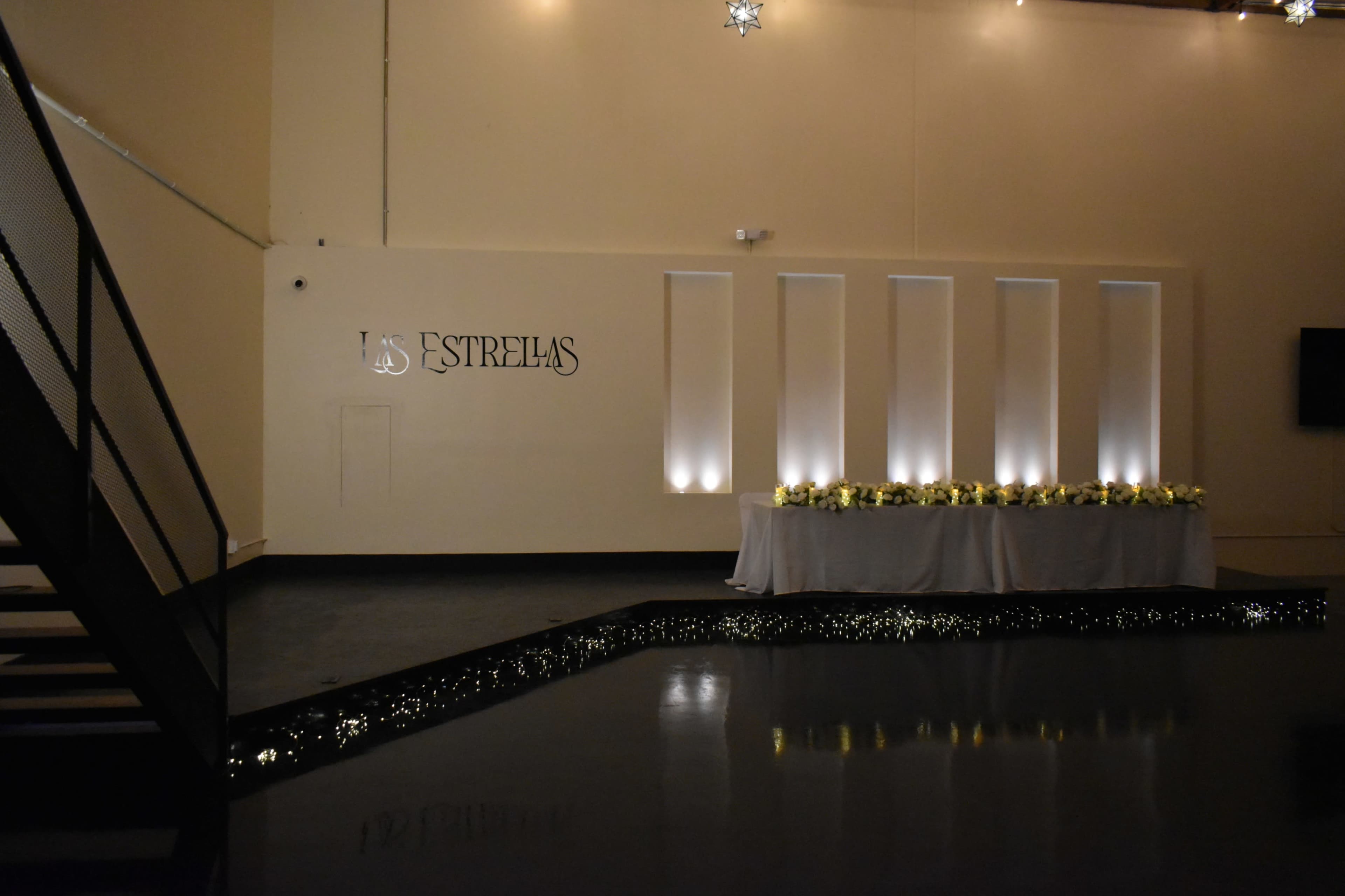 The image shows a decorated interior space featuring a wall with the name "LOS ESTRELLAS" and a illuminated table adorned with flowers and soft lighting.