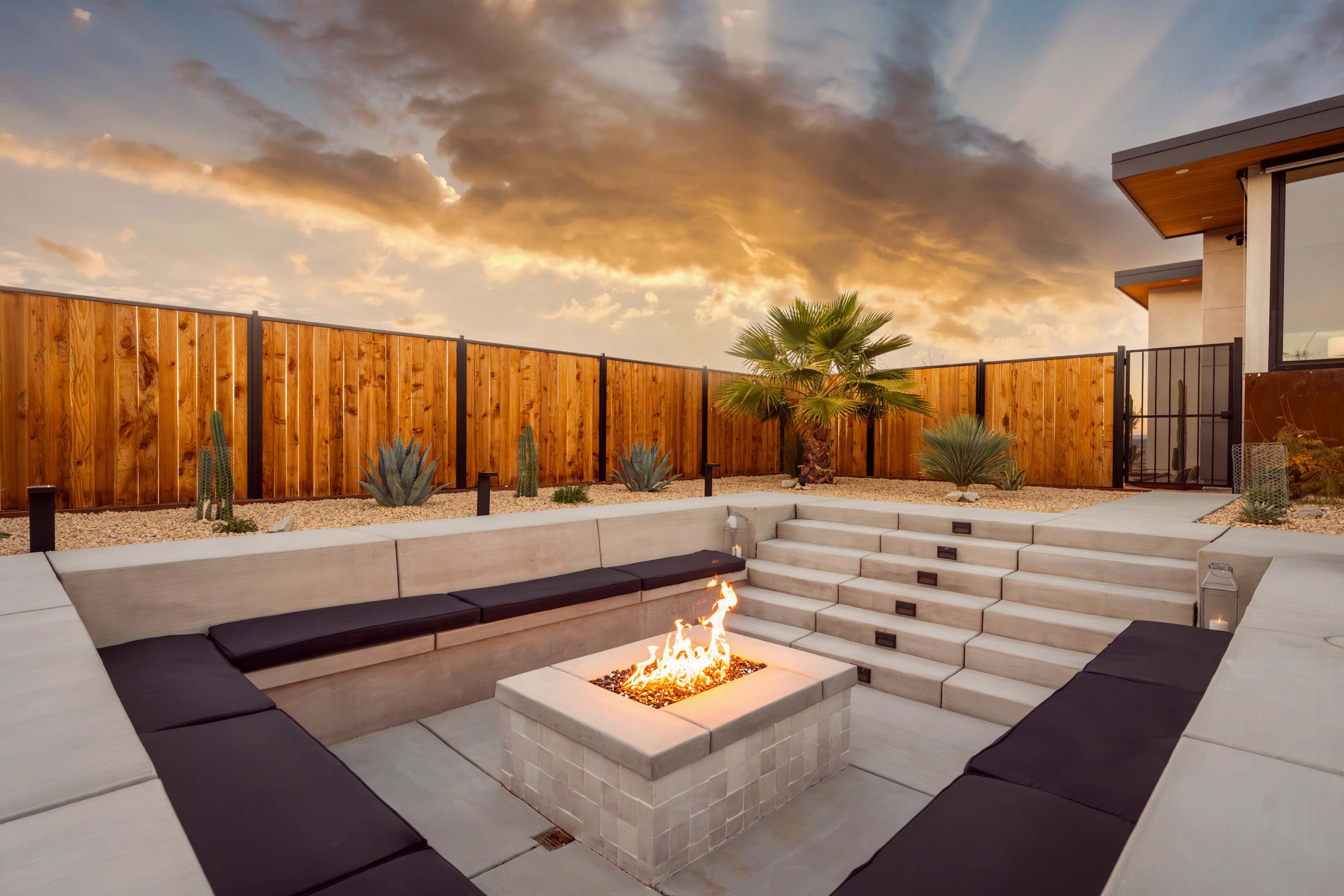 A modern outdoor patio features a fire pit surrounded by built-in seating, with a backdrop of tall wooden fencing and desert landscaping under a dramatic sky.