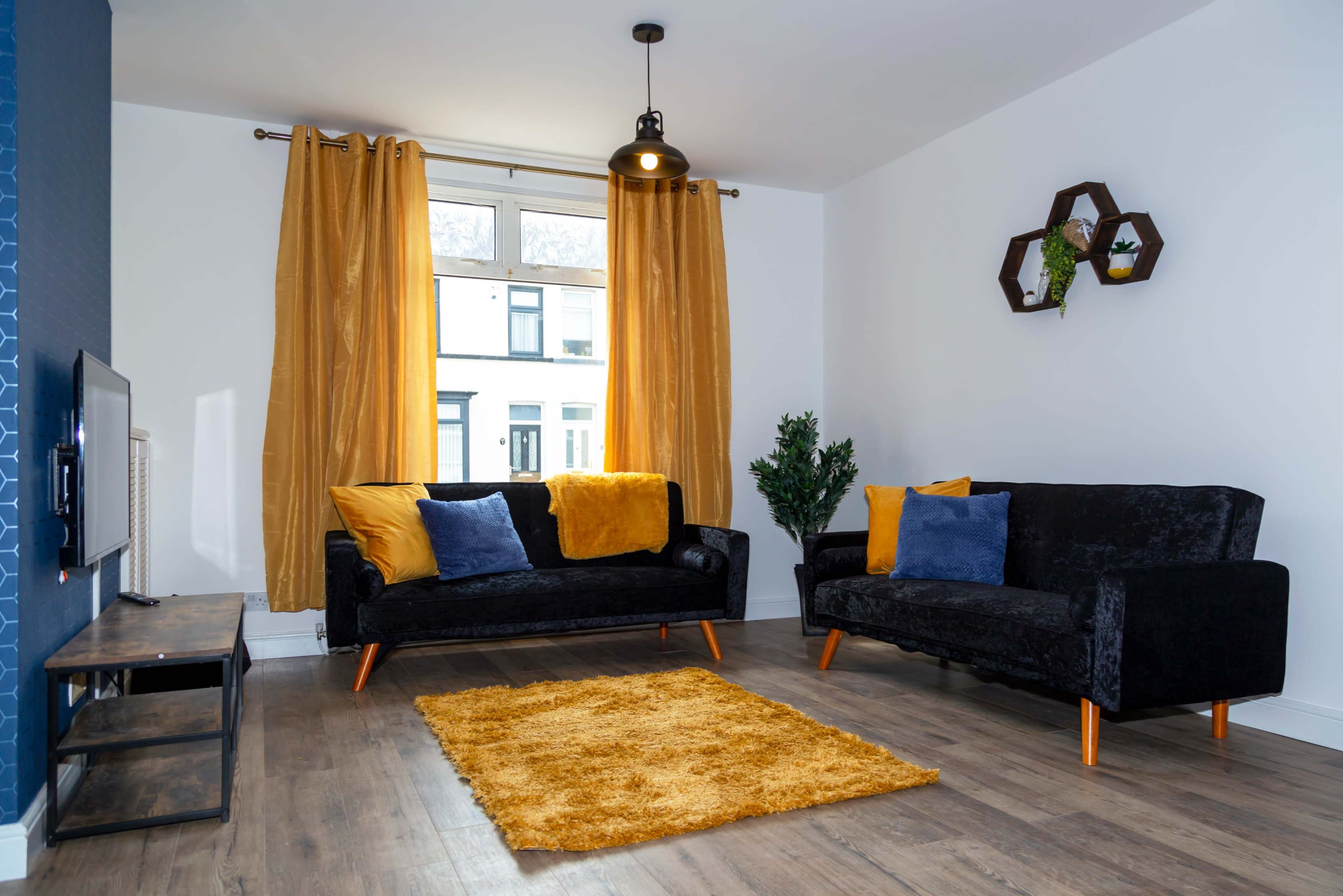 A modern living room features two black sofas with yellow and blue pillows, a yellow rug, and large windows dressed with mustard curtains.
