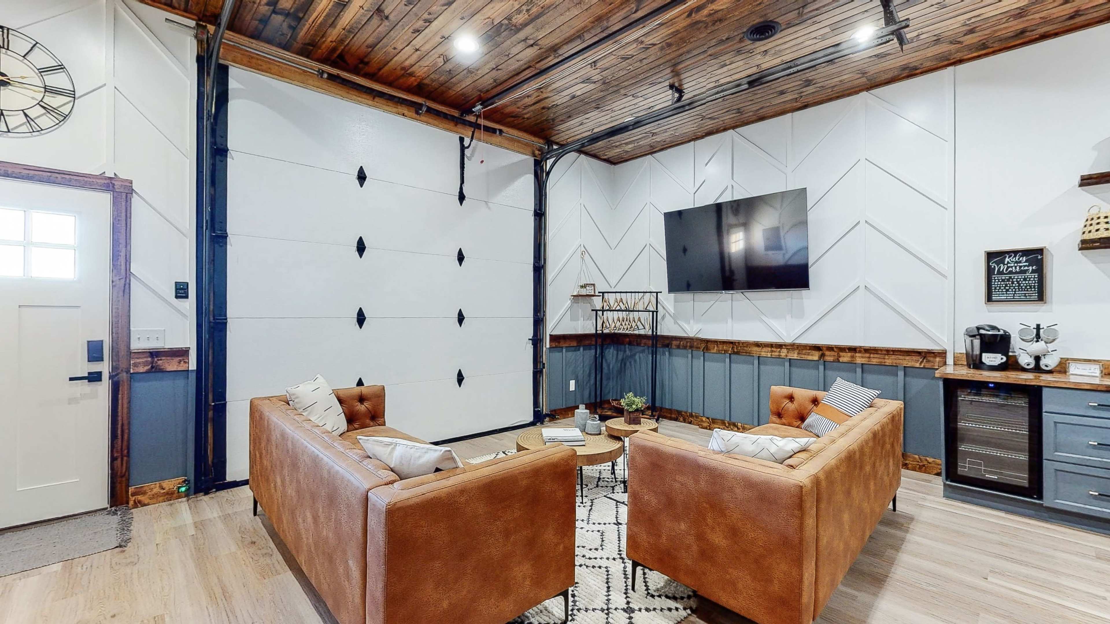 The room features two leather couches facing a wall-mounted television, with a wooden ceiling and a side counter displaying kitchen appliances.