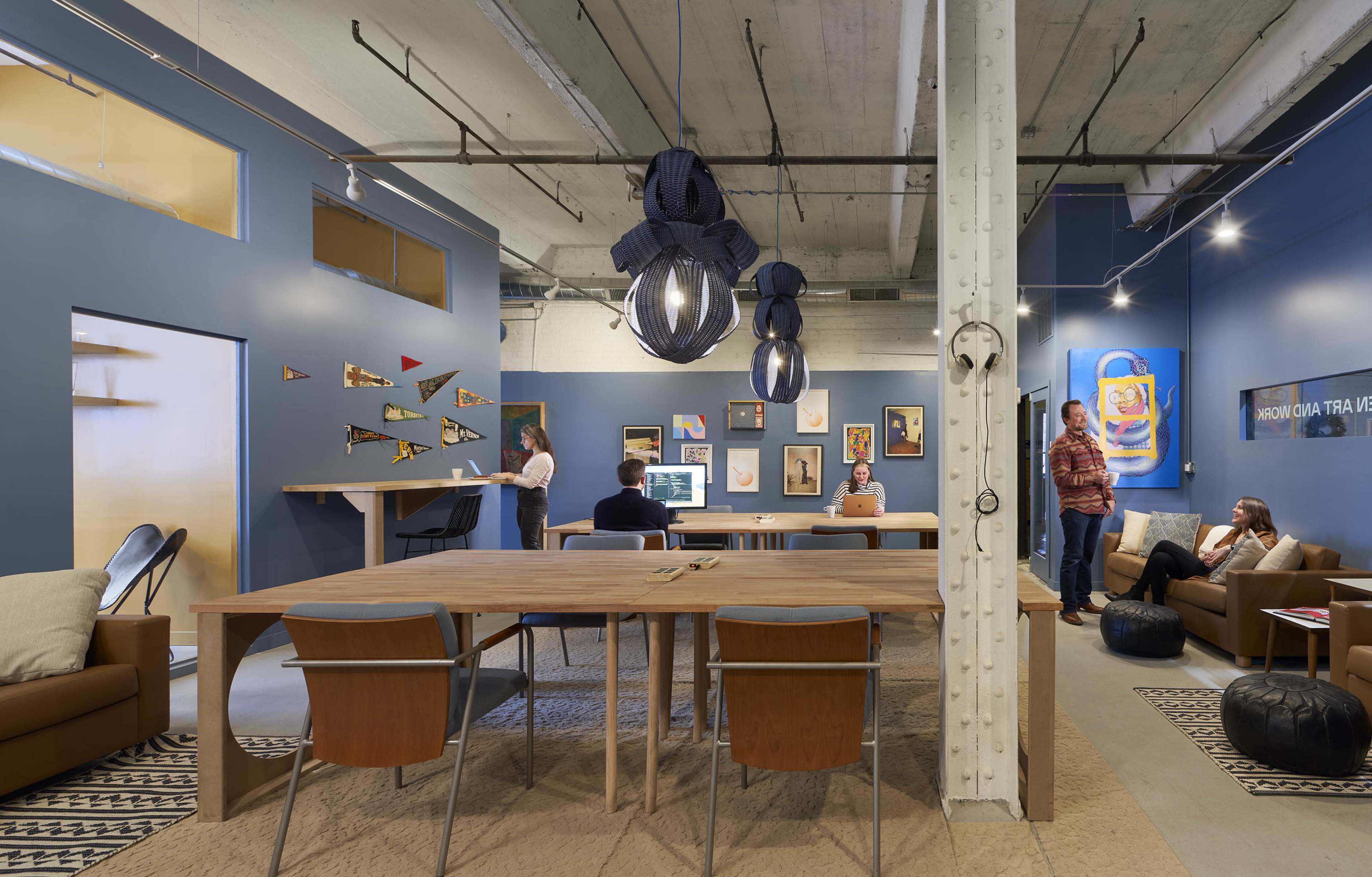 The image depicts a modern office space featuring a large wooden table, various seating arrangements, and colorful wall art in a blue-painted room.