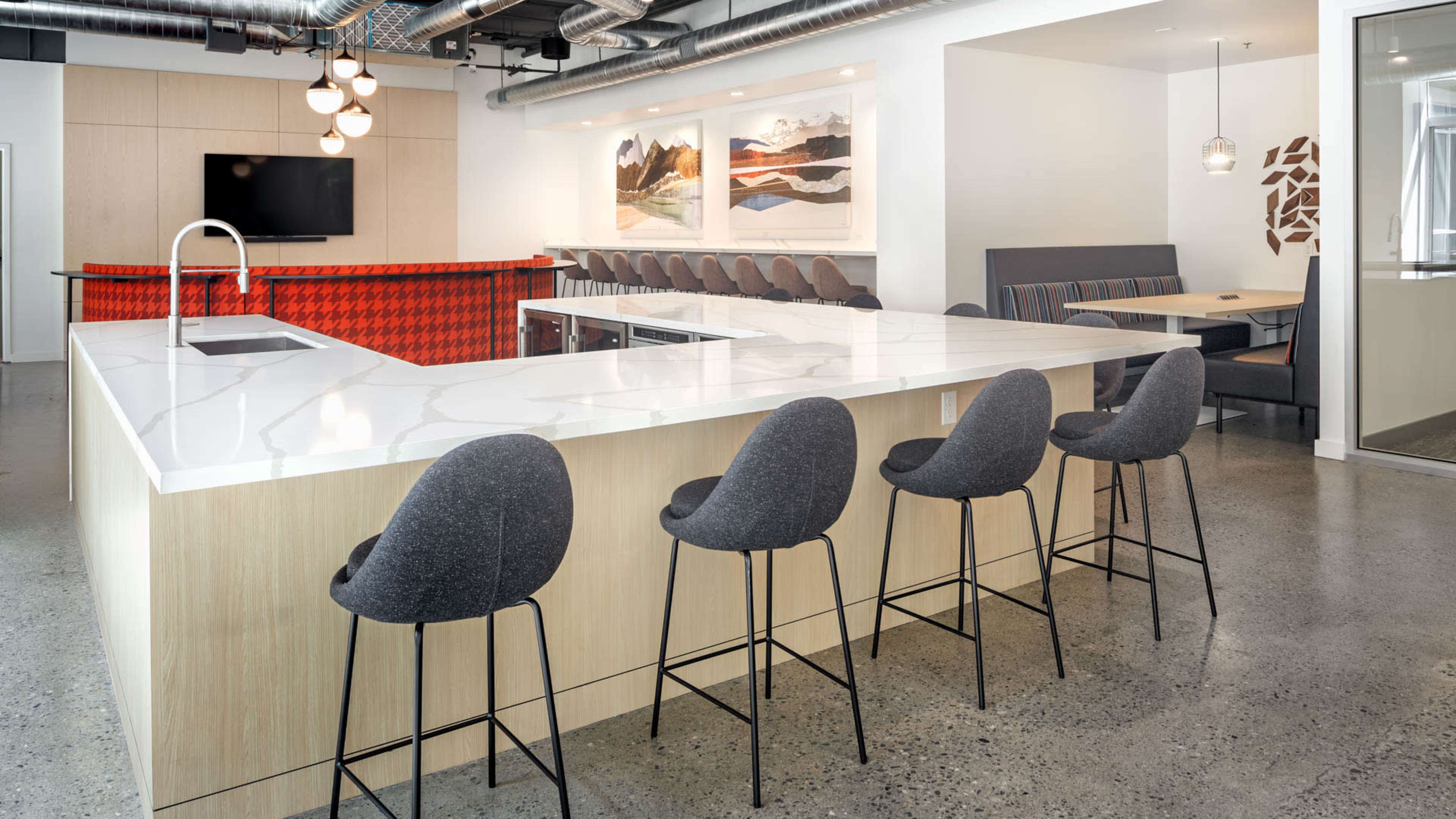 A modern kitchen area features a large white countertop with black barstools and a kitchenette in the background adorned with artwork.