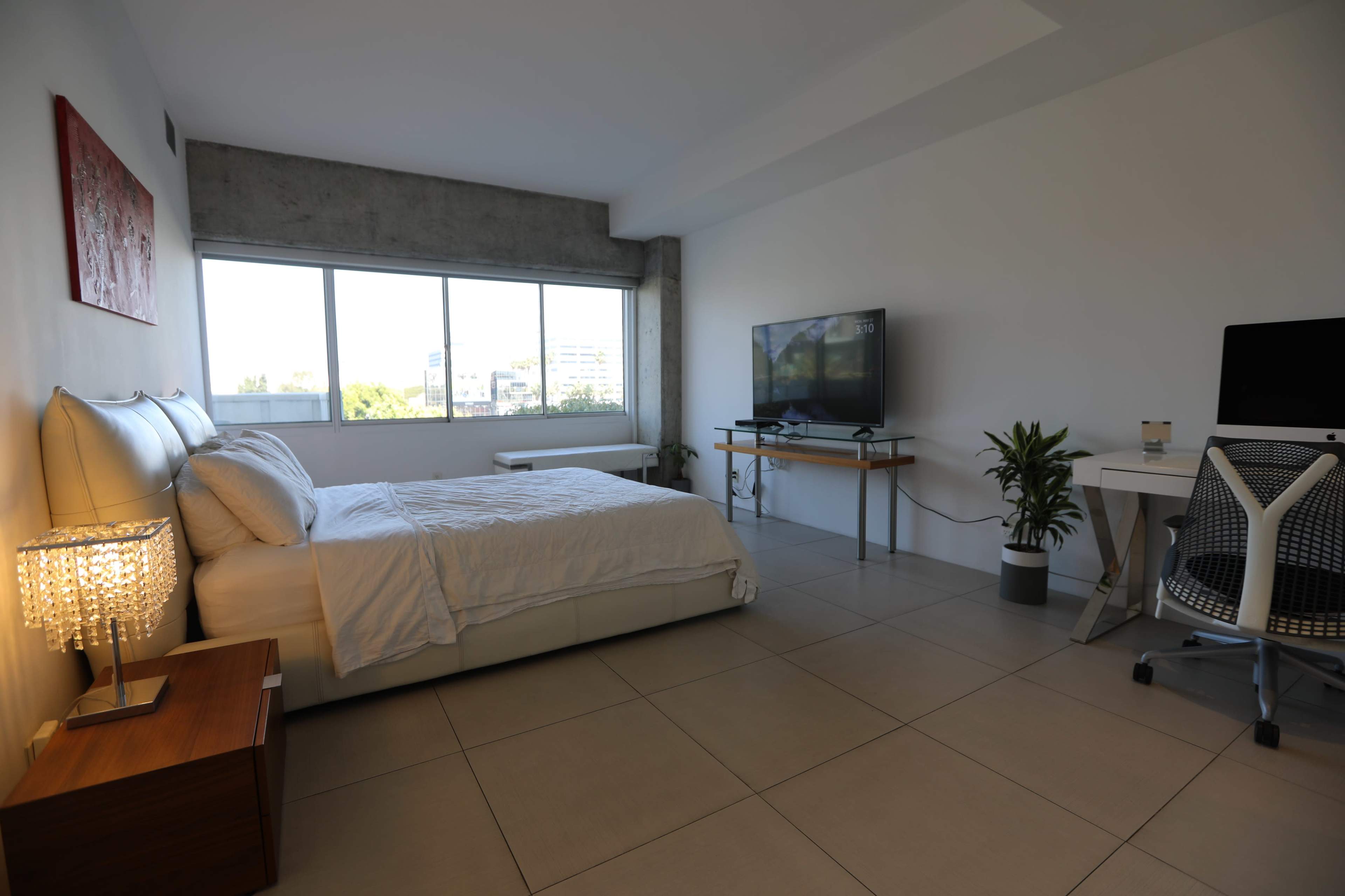 The image shows a modern bedroom featuring a bed, a television, a work desk, and large windows allowing natural light.