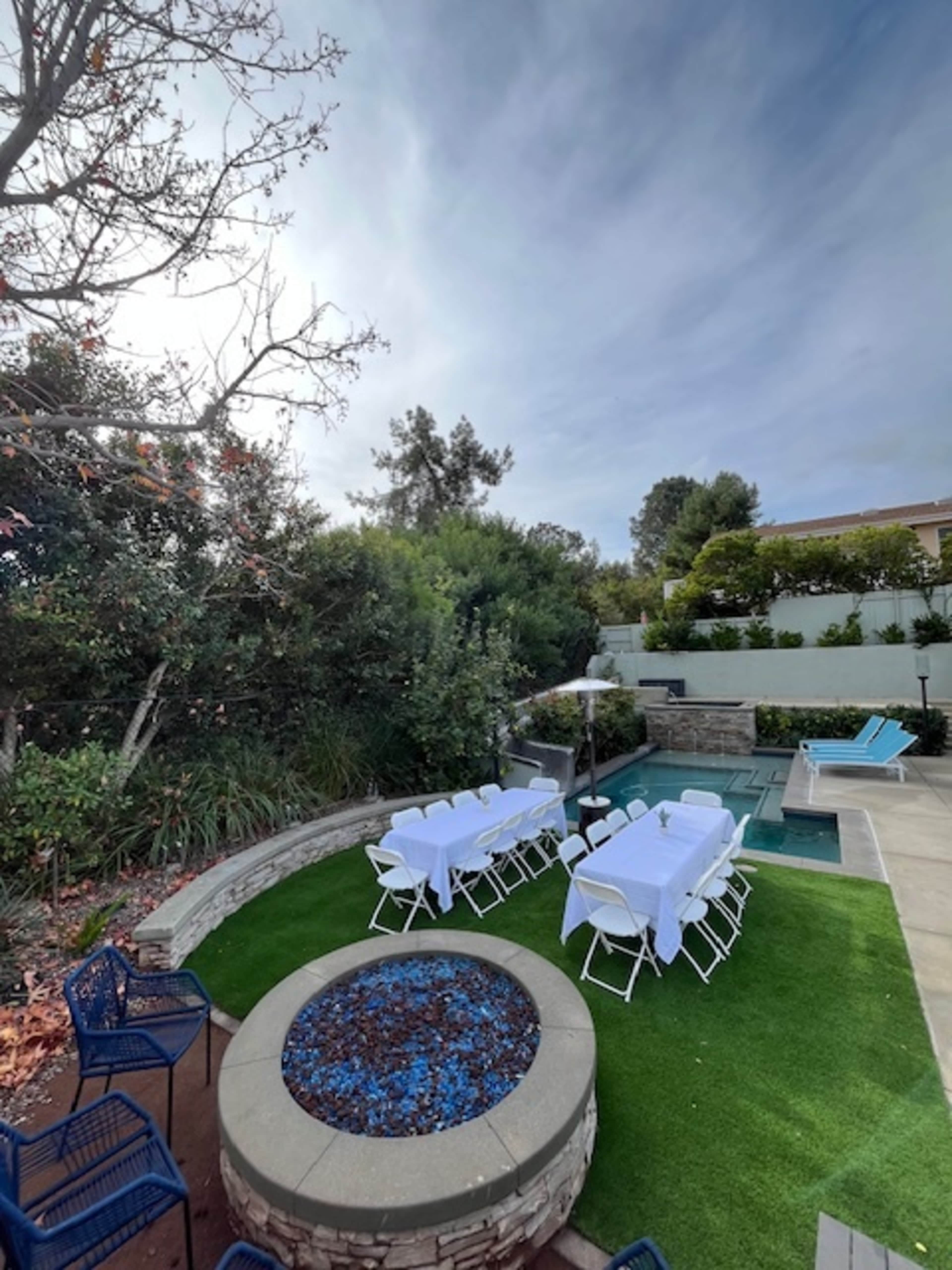 A neatly arranged outdoor seating area with white tables and chairs is set up near a pool and surrounded by greenery.