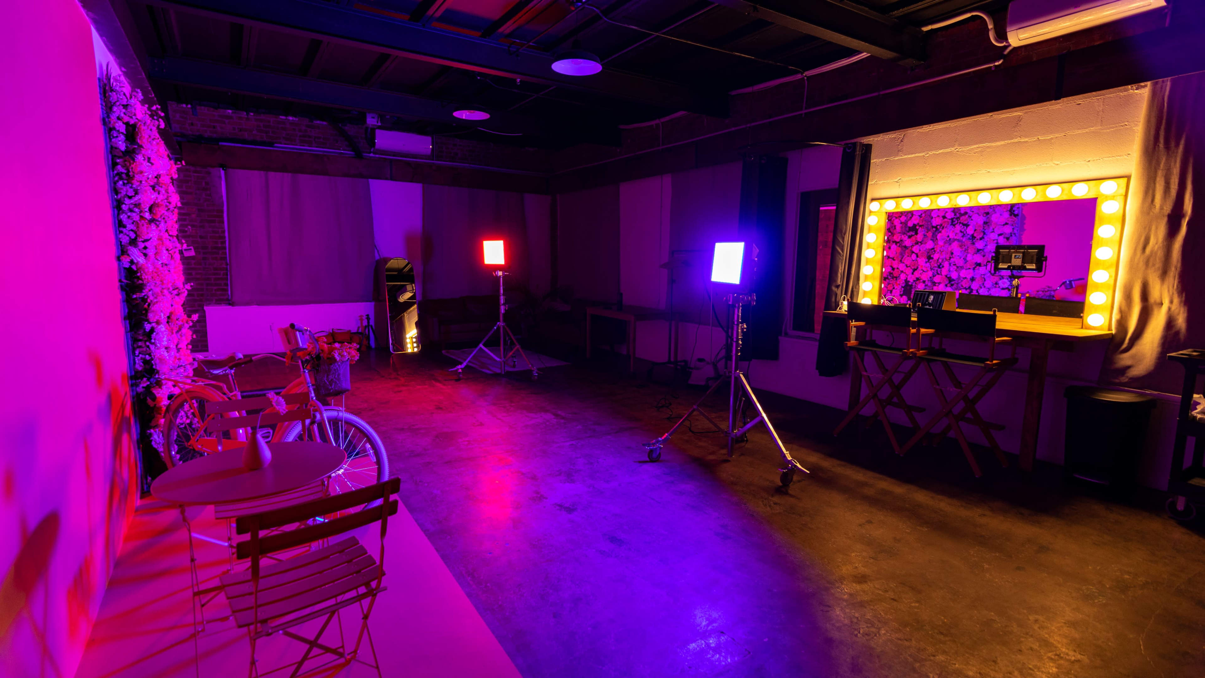 A dimly lit studio space with colorful lighting, a mirror with light bulbs, and equipment set up for a photoshoot.