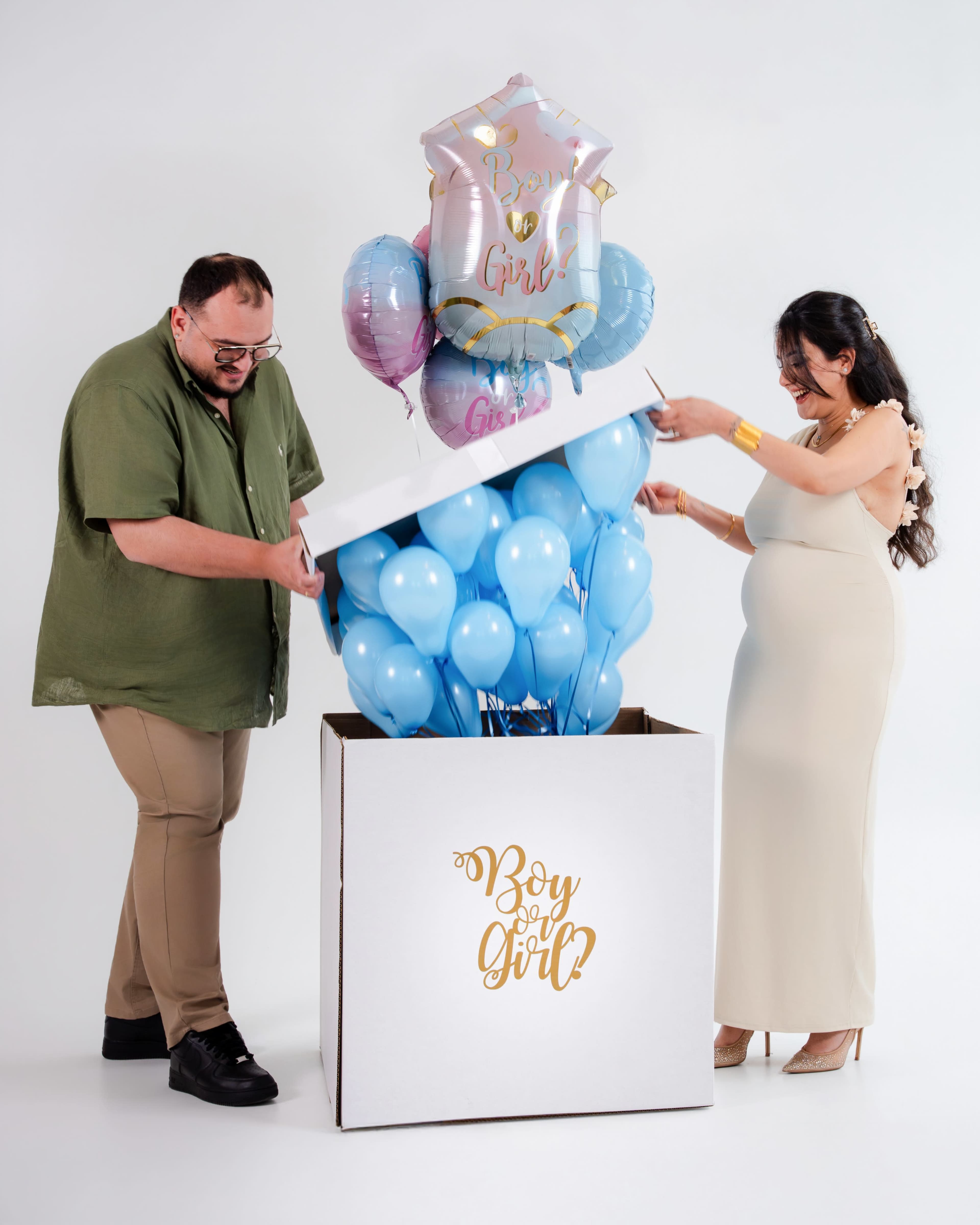 A couple is opening a large gift box filled with blue balloons and decorated with pink and blue balloons on top, indicating a gender reveal event.