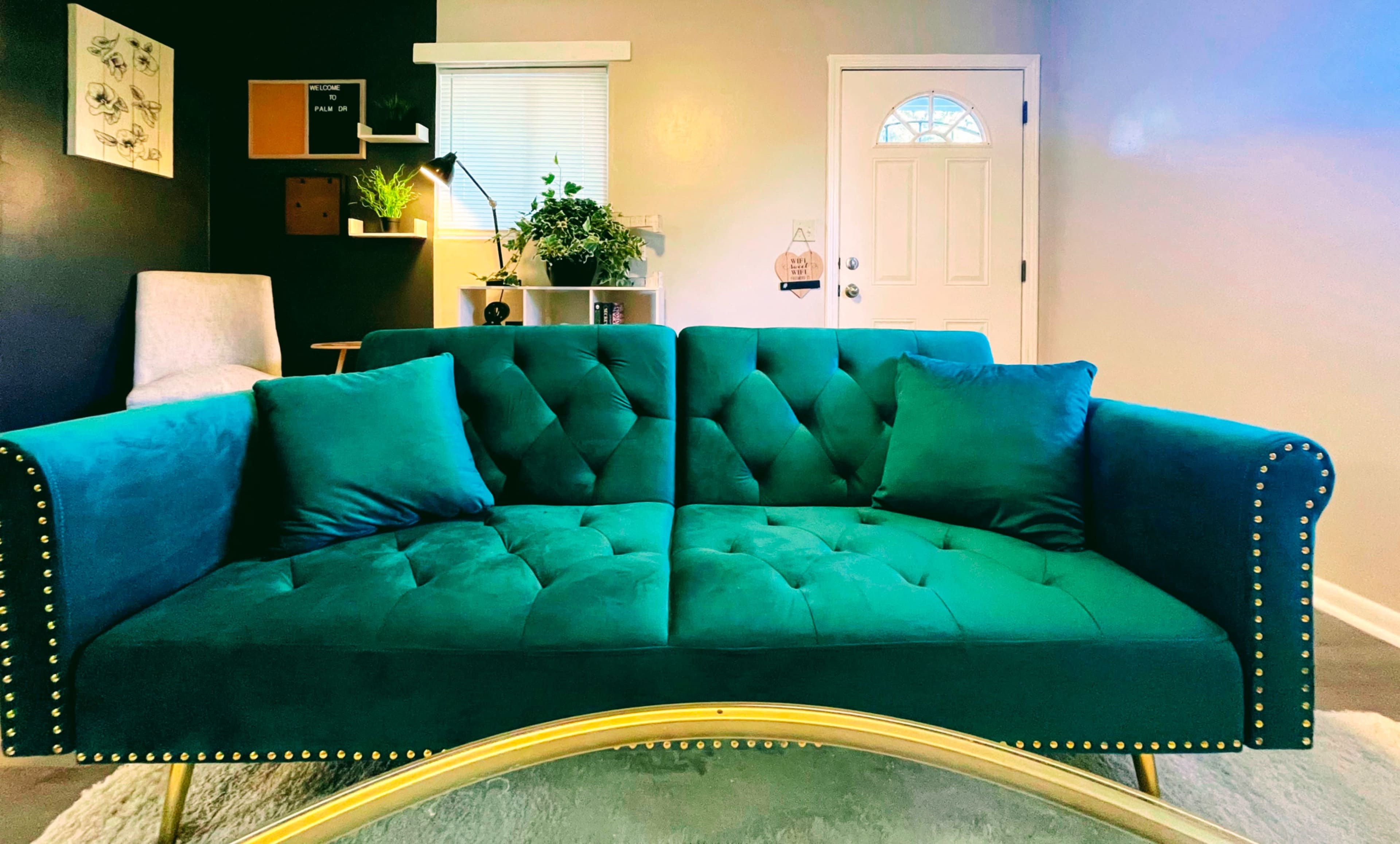 A teal tufted sofa with golden studs, placed in a well-lit room featuring a door and plants.