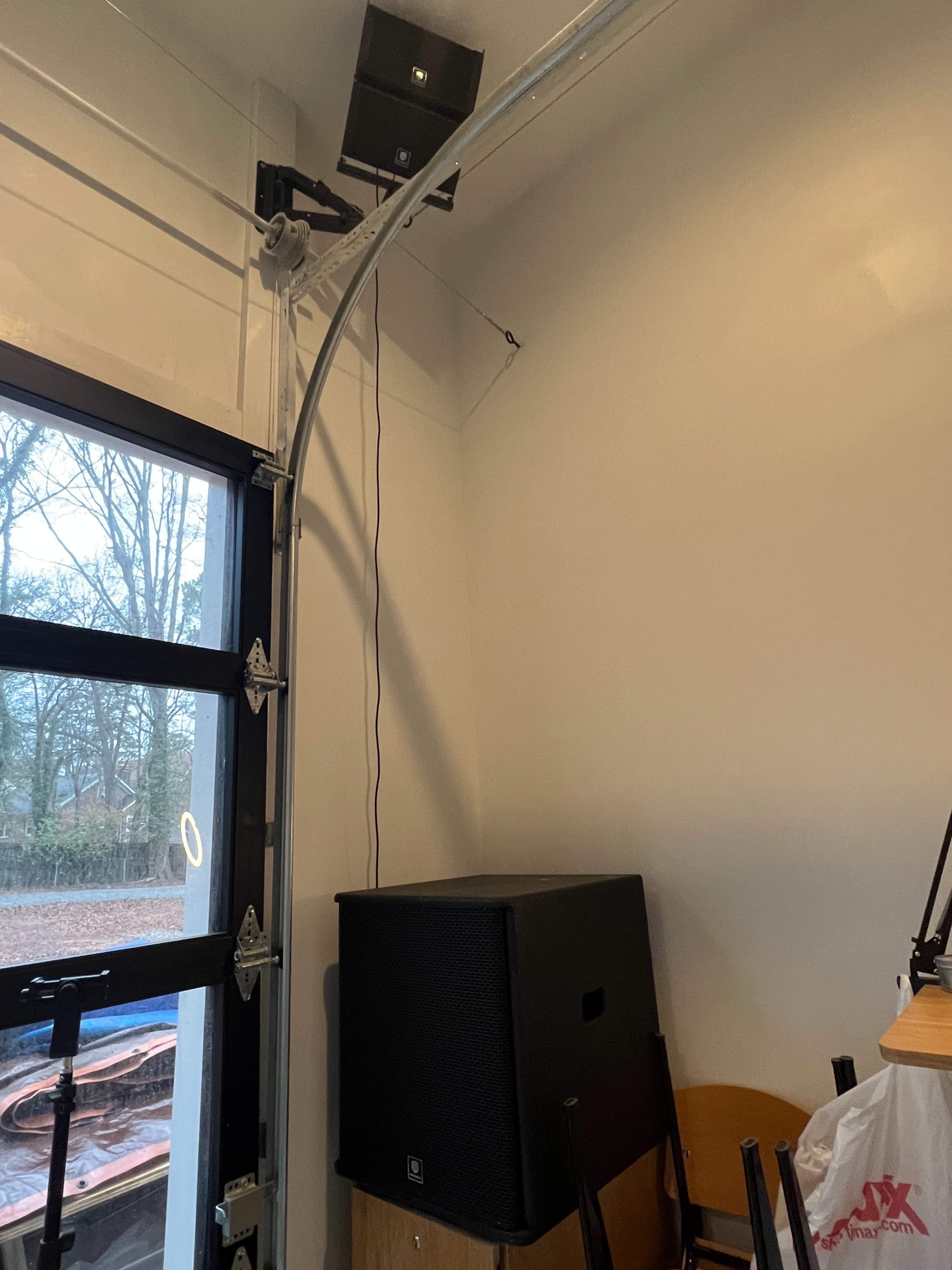A corner of a room with a black speaker on a wooden surface, a mounted speaker above, and a partially open garage door revealing an outdoor view.