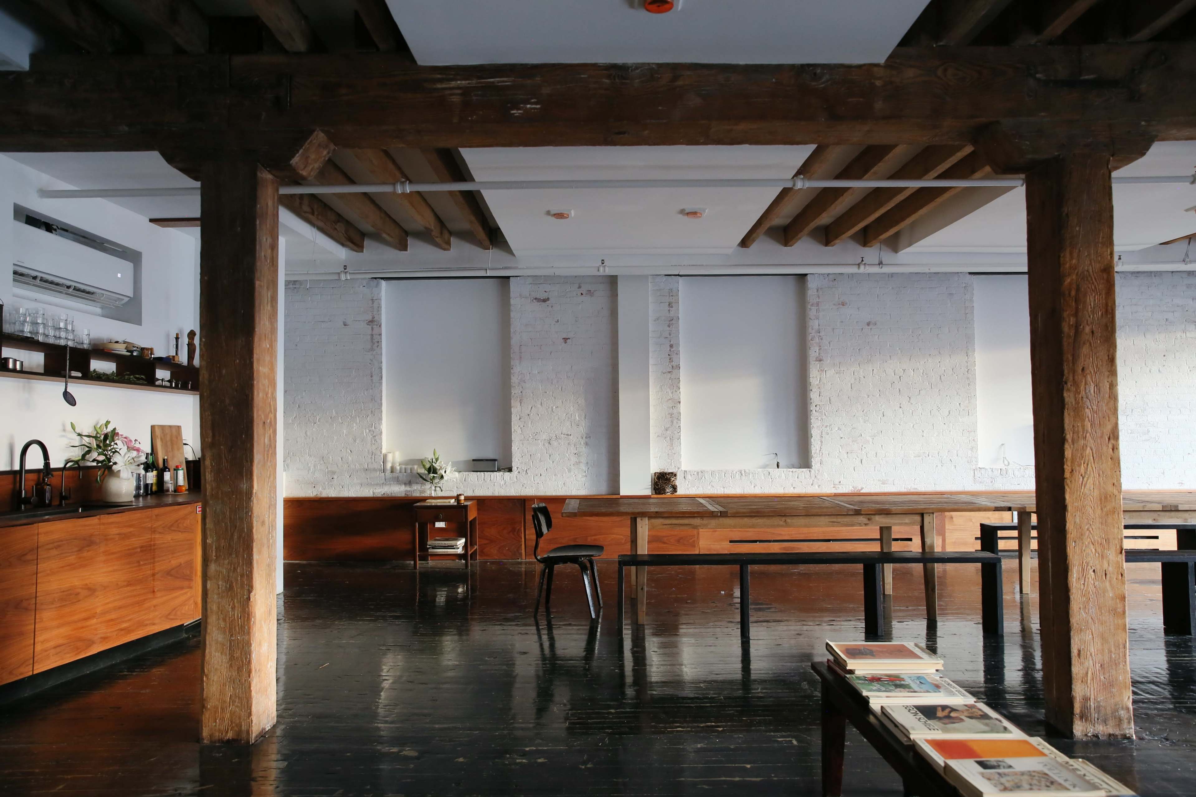 The image shows a spacious interior with exposed wooden beams, dark polished floors, and a minimalist kitchen area featuring wooden cabinetry and a long communal table.