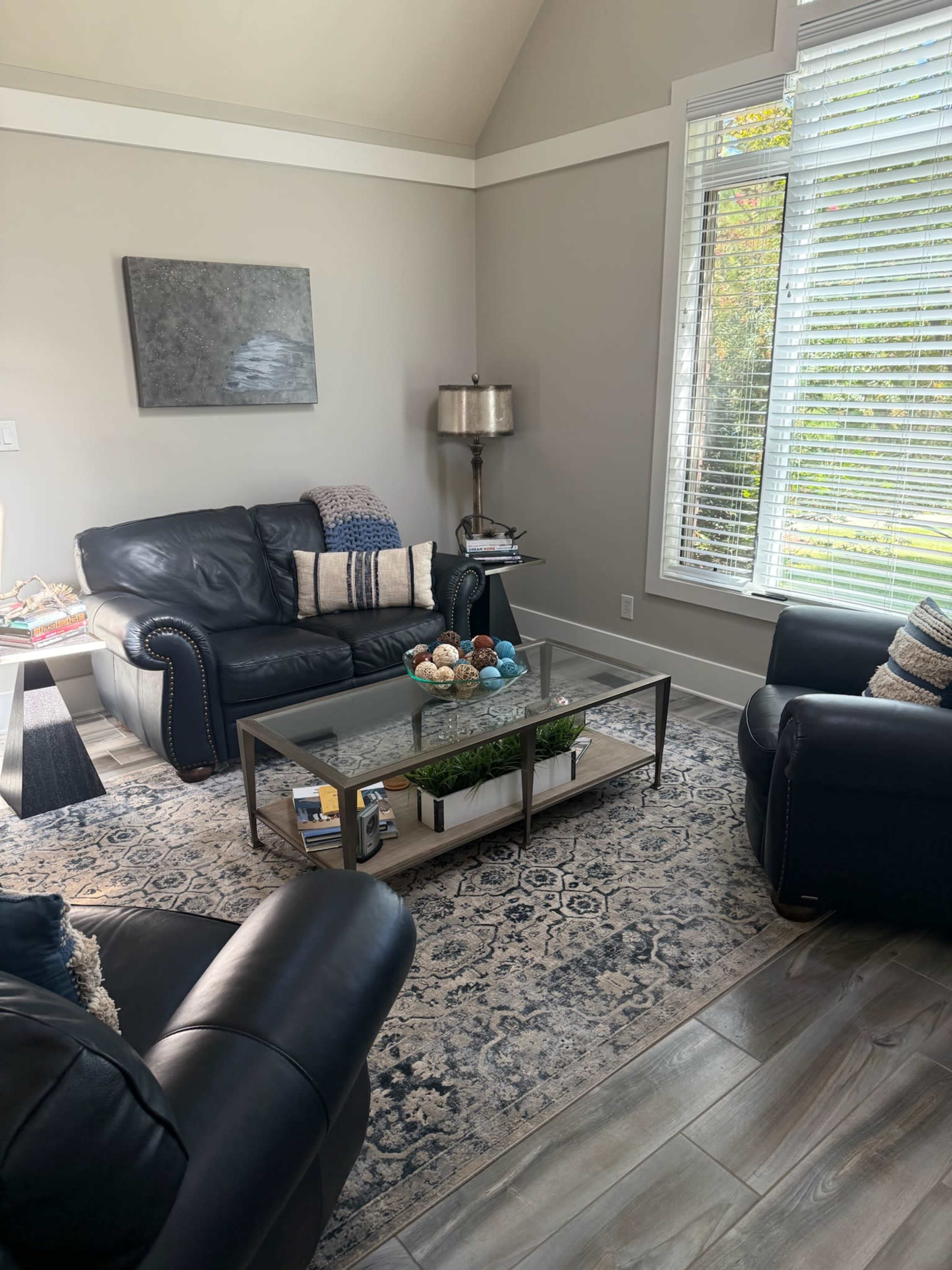 The scene shows a living room with black leather sofas, a glass coffee table, and large windows allowing natural light to illuminate the space.